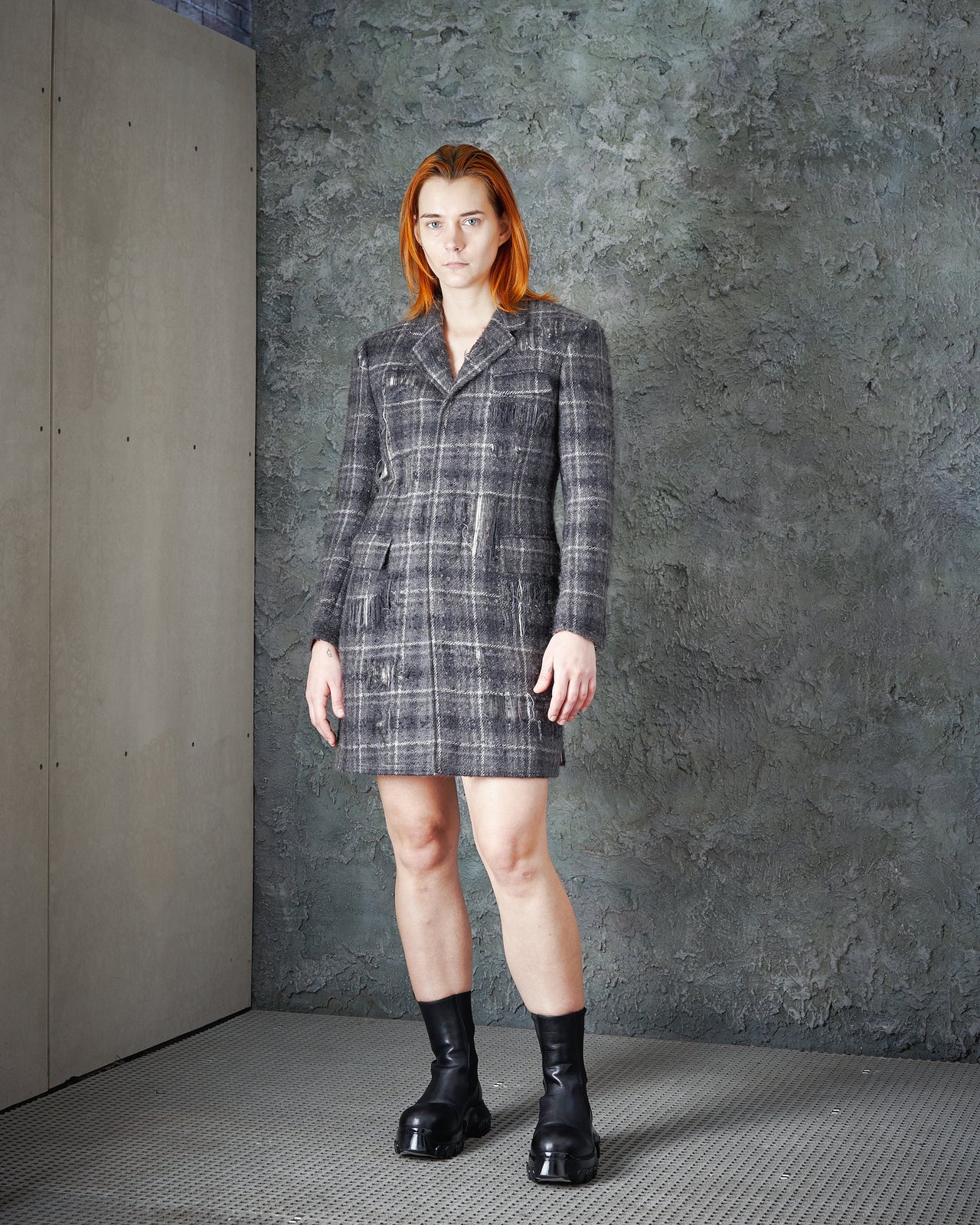 A person with straight red hair stands indoors against a textured gray wall, wearing the FW 2016 Distressed Tartan Mohair Coat by Thom Browne as a grey plaid blazer dress paired with black chunky boots.