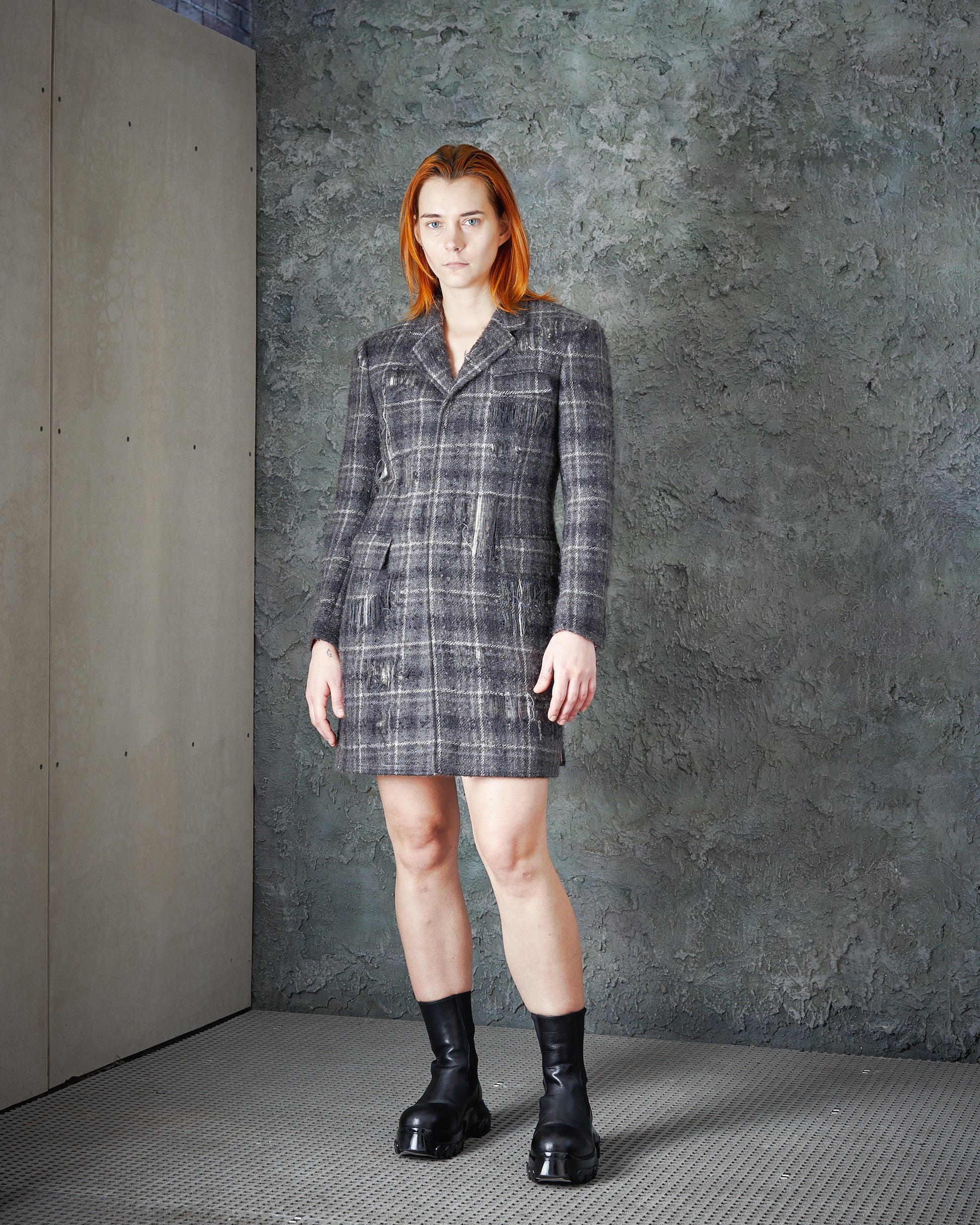 A person with straight red hair stands indoors against a textured gray wall, wearing the FW 2016 Distressed Tartan Mohair Coat by Thom Browne as a grey plaid blazer dress paired with black chunky boots.