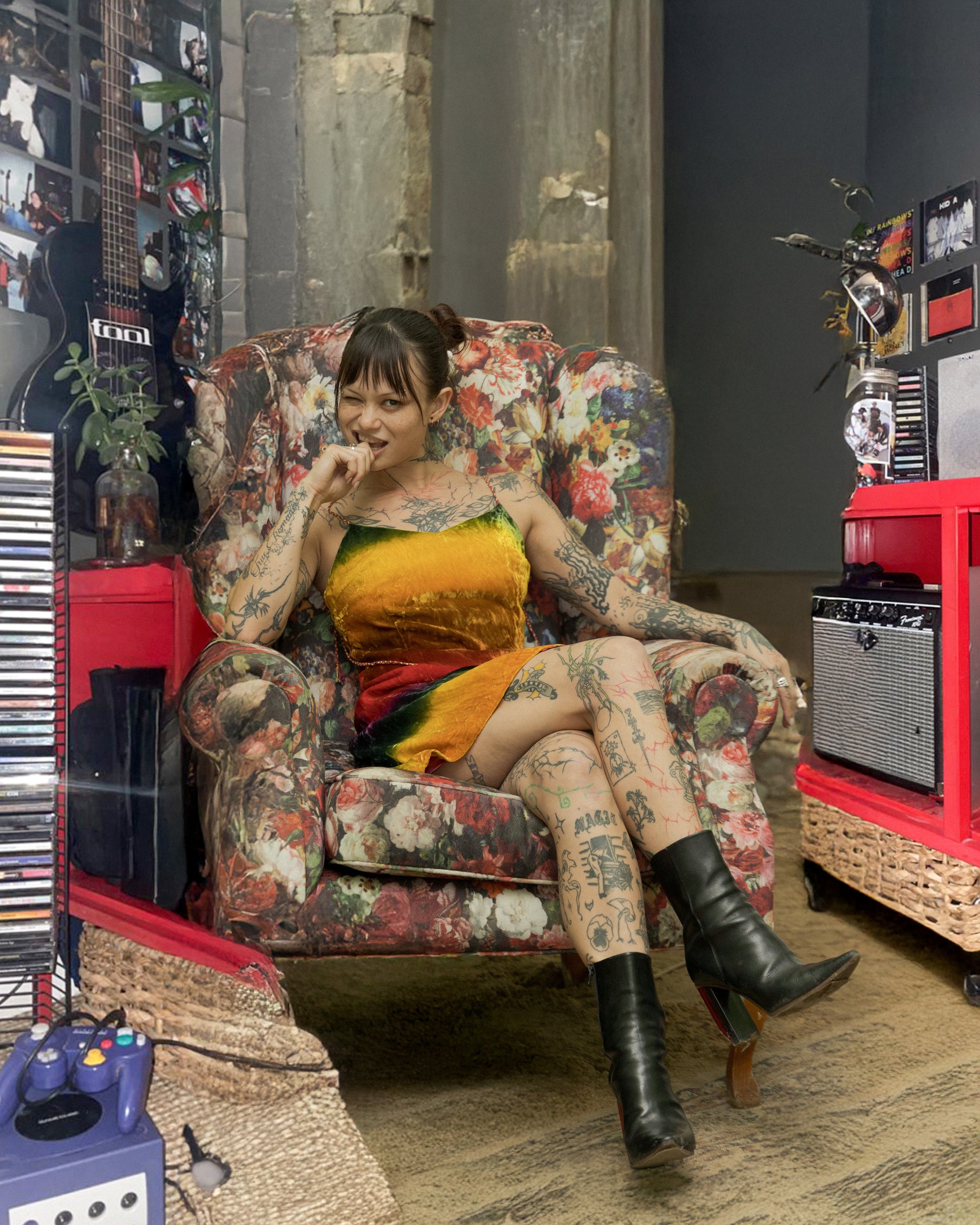 Person with tattoos sitting on a floral-patterned chair in a room with musical equipment wearing a multicoloured Todd Oldham mini dress
