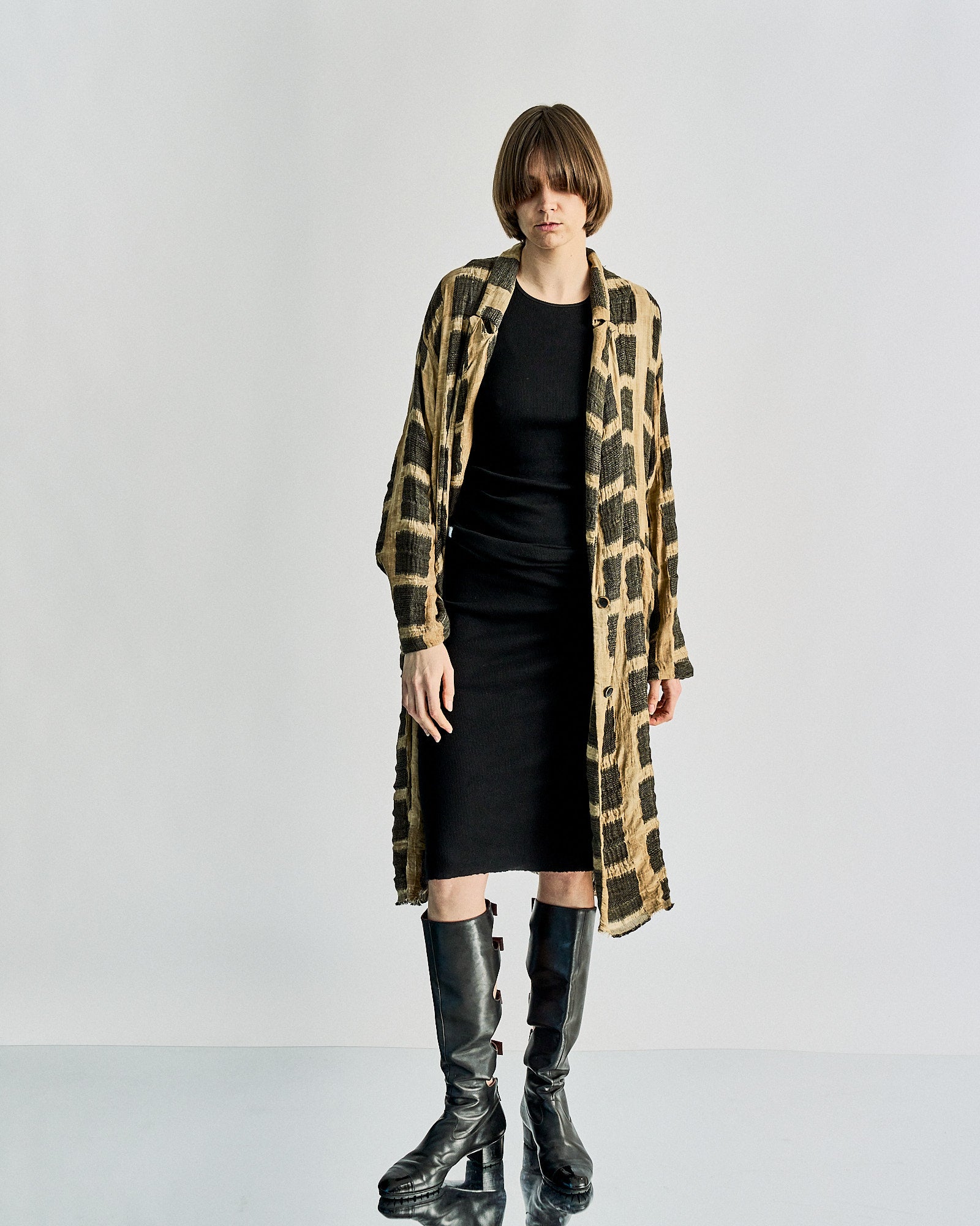 A person stands against a light grey background wearing a long black dress, knee-high black boots, and a tan Uma Wang coat with a dark square pattern. The expression is neutral.
