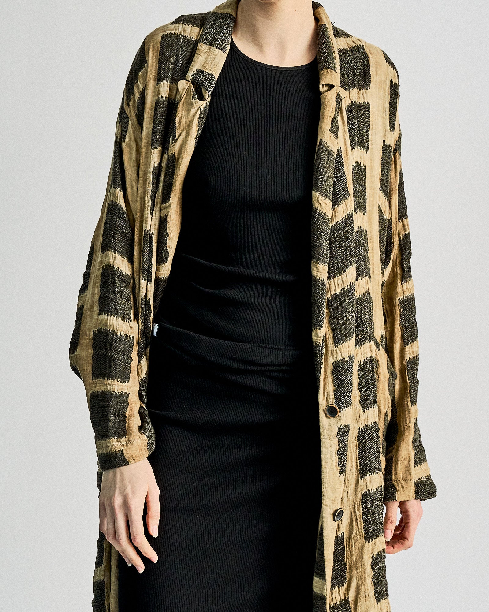 A person wearing a long, tan Uma Wang coat with large black checkered patterns over a fitted black dress. The outfit exudes a stylish, modern aesthetic.