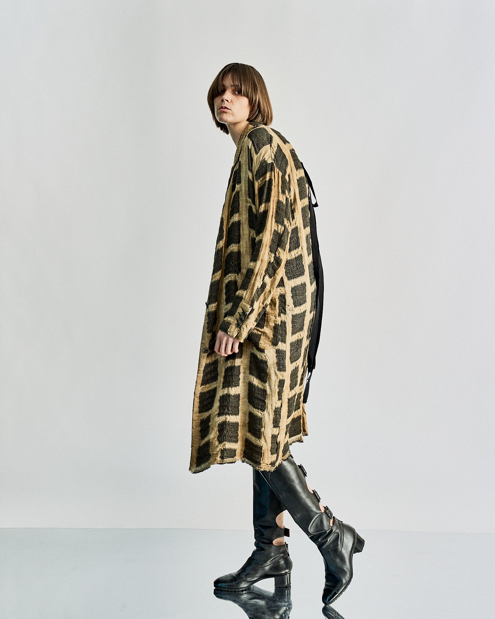 Person with a bob haircut wears a long, checkered Uma Wang coat and black knee-high boots, walking confidently on a reflective surface.