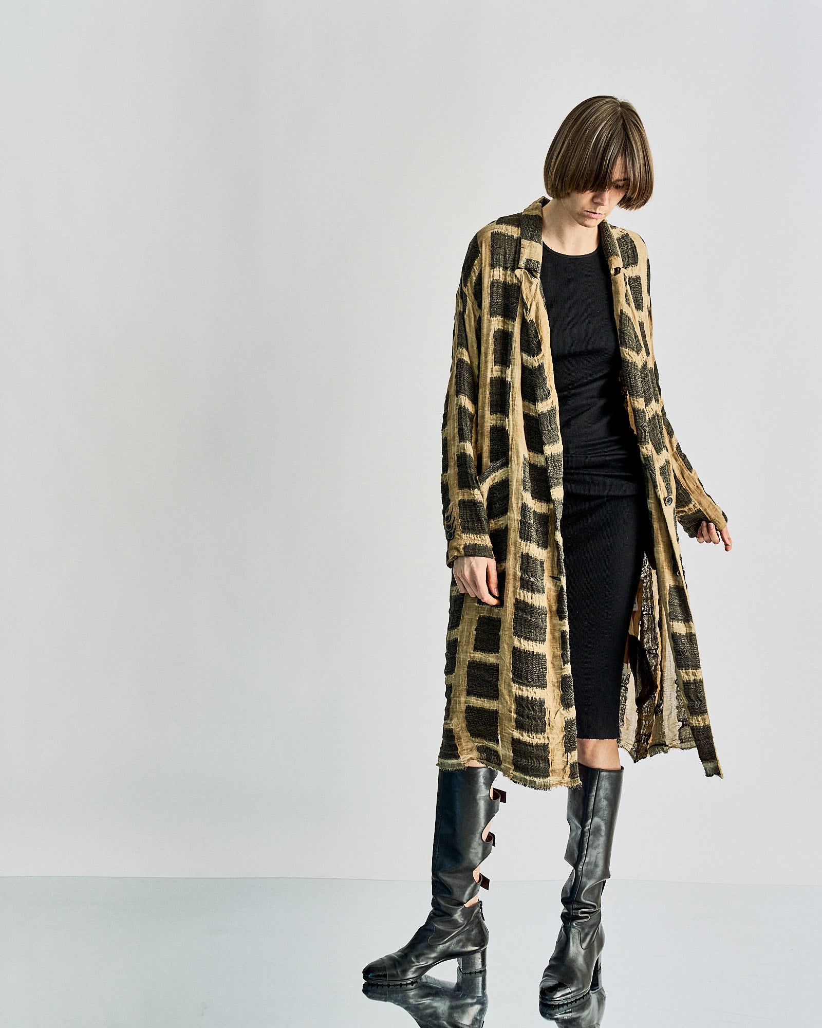 A person wearing a black dress, long plaid Uma Wang coat, and knee-high black boots stands against a plain background, exuding a stylish and contemplative tone.