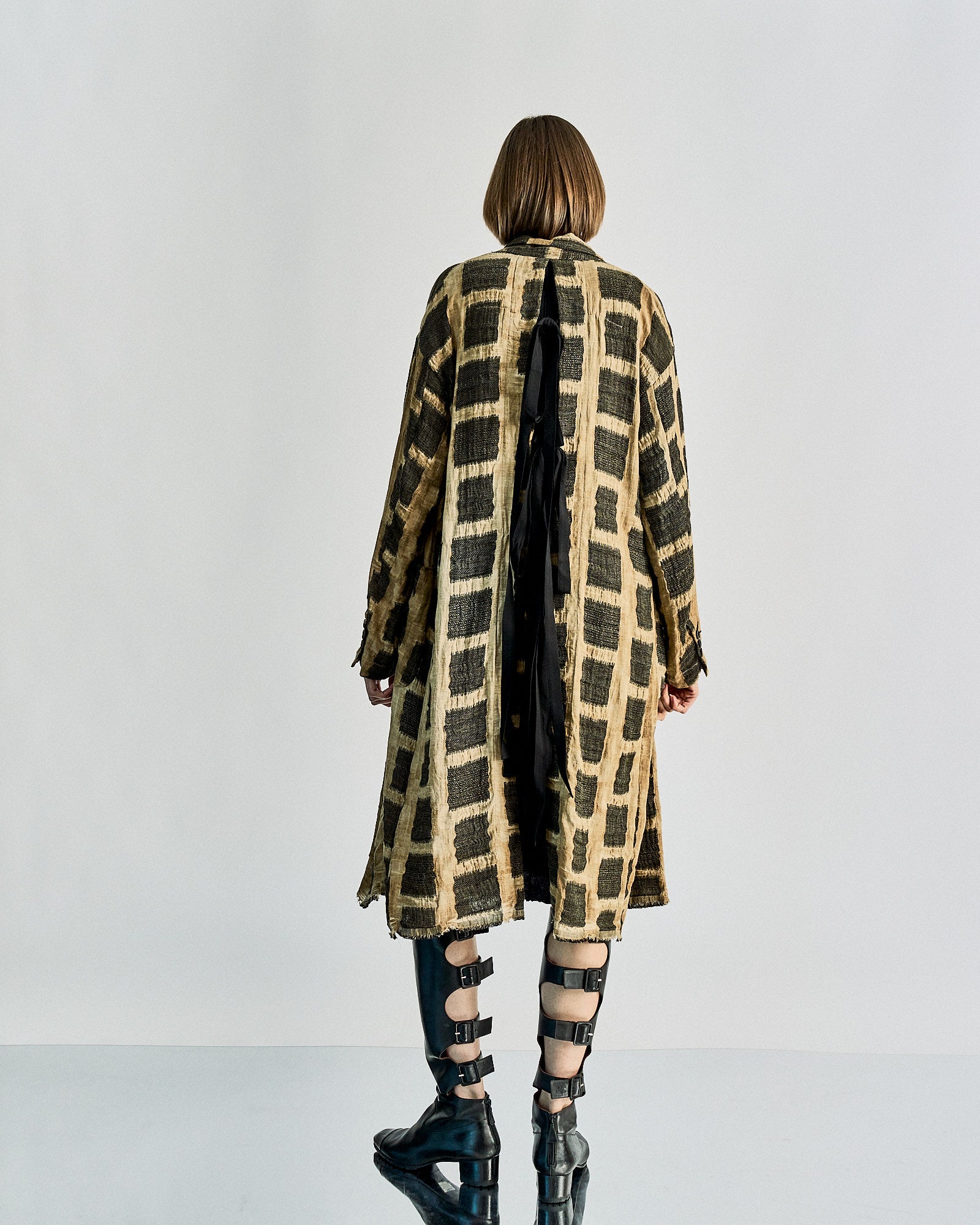 Person with short hair in a long, textured Uma Wang coat with a black and beige grid pattern, facing away. They wear tall, strappy black boots