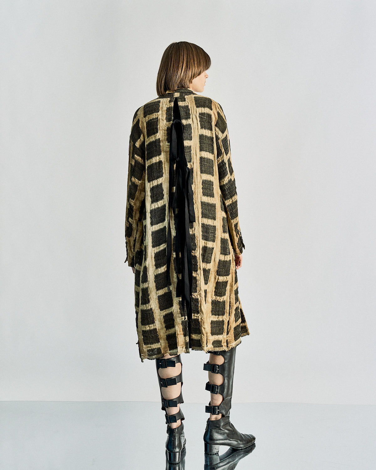 A longline Uma Wang coat in a textural linen with a black and white animal pattern, featuring a slim sculptural collar, two-button front, and pockets at the hips. The coat is on a mannequin and the model is wearing a black dress and black boots.