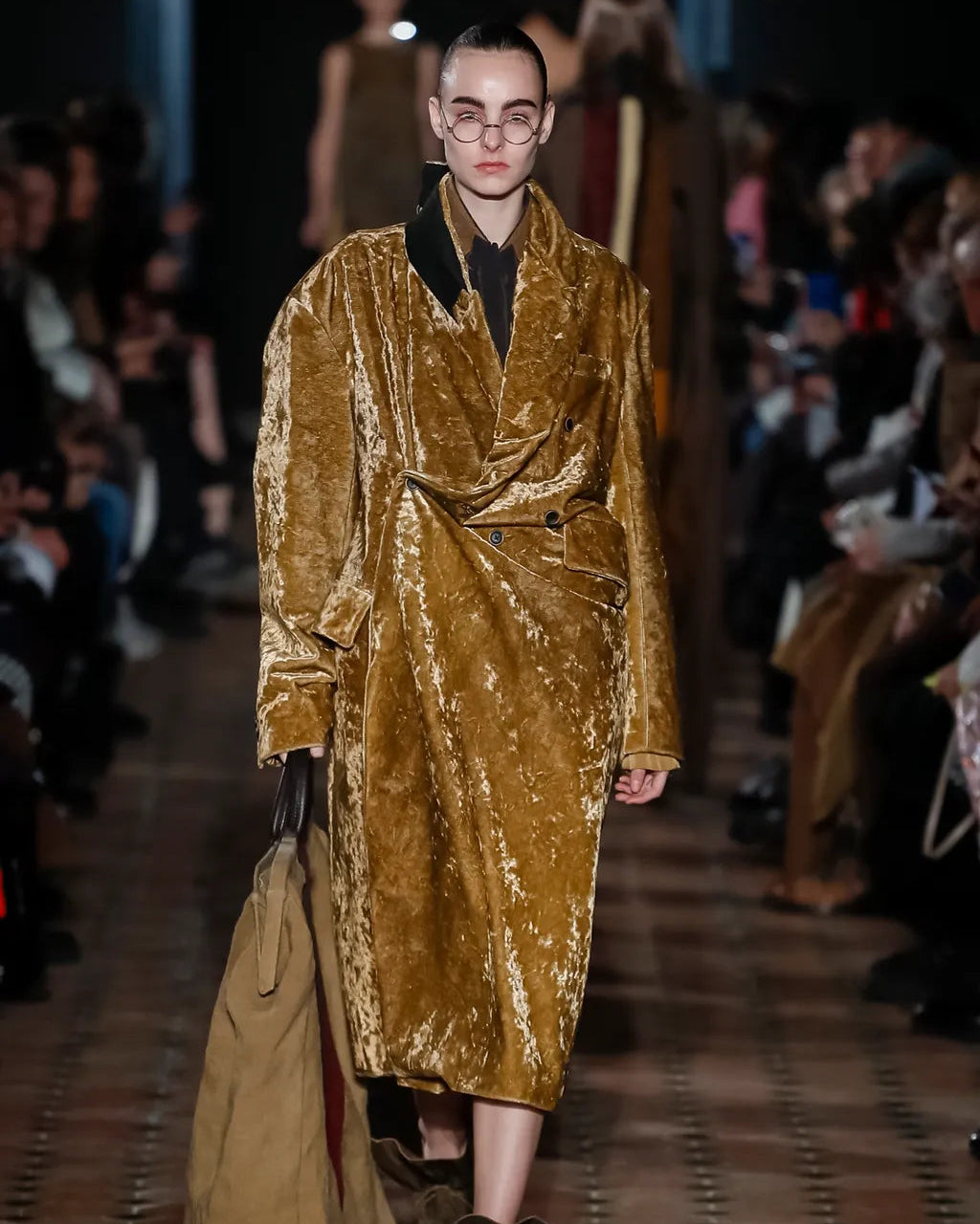 Model wearing a gold velvet Uma Wang coat on a runway with an audience.