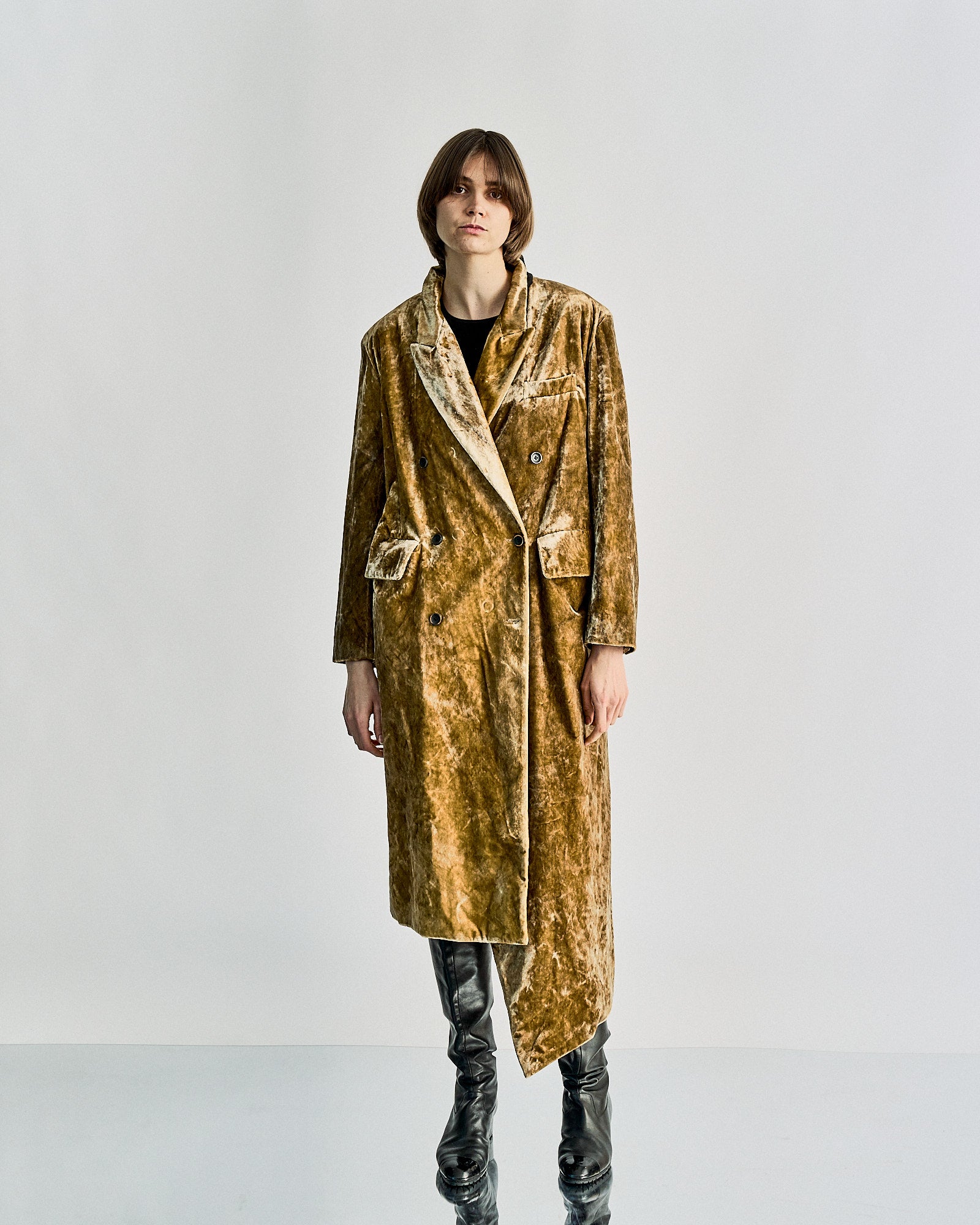 Person wearing a long, velvet gold Uma Wang coat with a slightly asymmetrical hem and black leather boots. The expression is neutral, set against a plain white background.