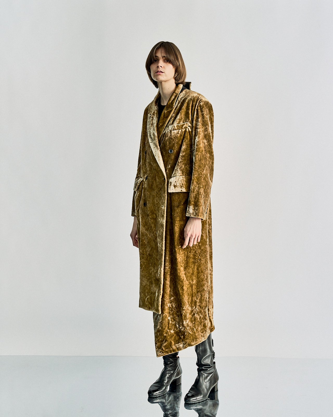 Person wearing a long, velvet gold Uma Wang coat with a slightly asymmetrical hem and black leather boots. The expression is neutral, set against a plain white background.