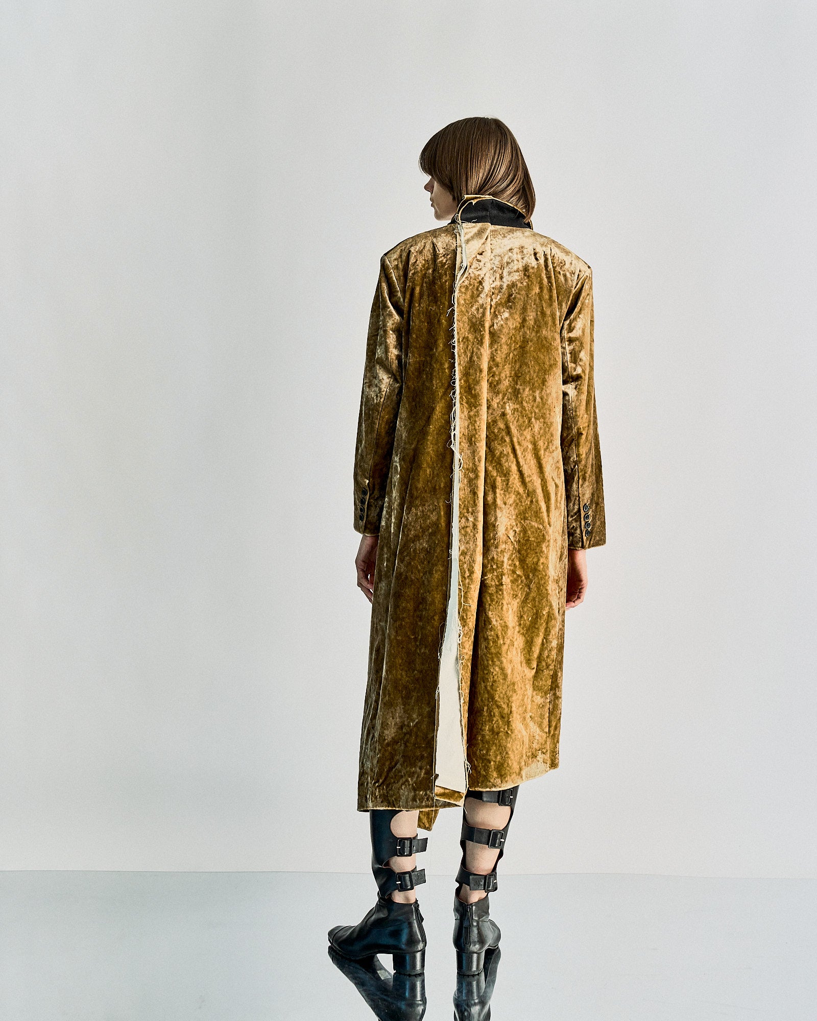 A person with short hair stands facing away, wearing a long velvet Uma Wang coat with a raw edge detail and black buckle boots. 