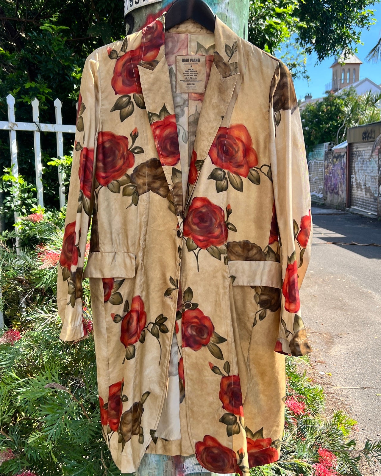 Floral-patterned Uma Wang coat hanging on a wooden post with greenery and a building in the background