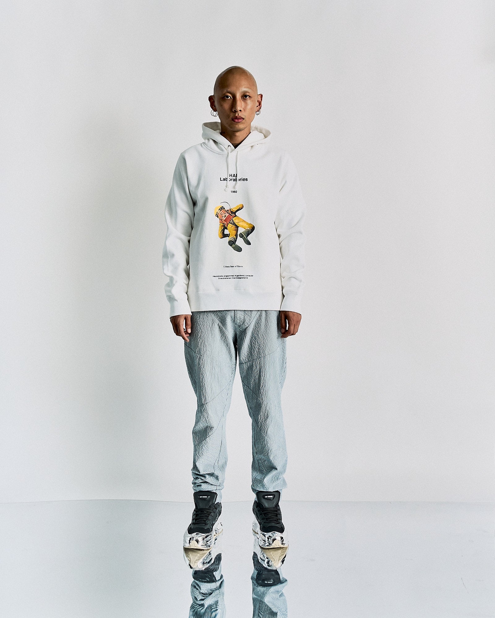 A person standing in a studio setting, wearing a white Undercover hoodie with a vintage-inspired 2001: A Space Odyssey print and gray pants.