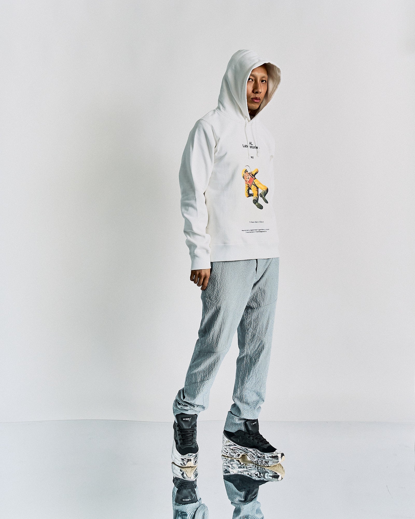 A person stands against a plain background wearing a white Undercover hoodie with a cartoon graphic, gray pants, and chunky sneakers. The tone is modern and minimalistic.