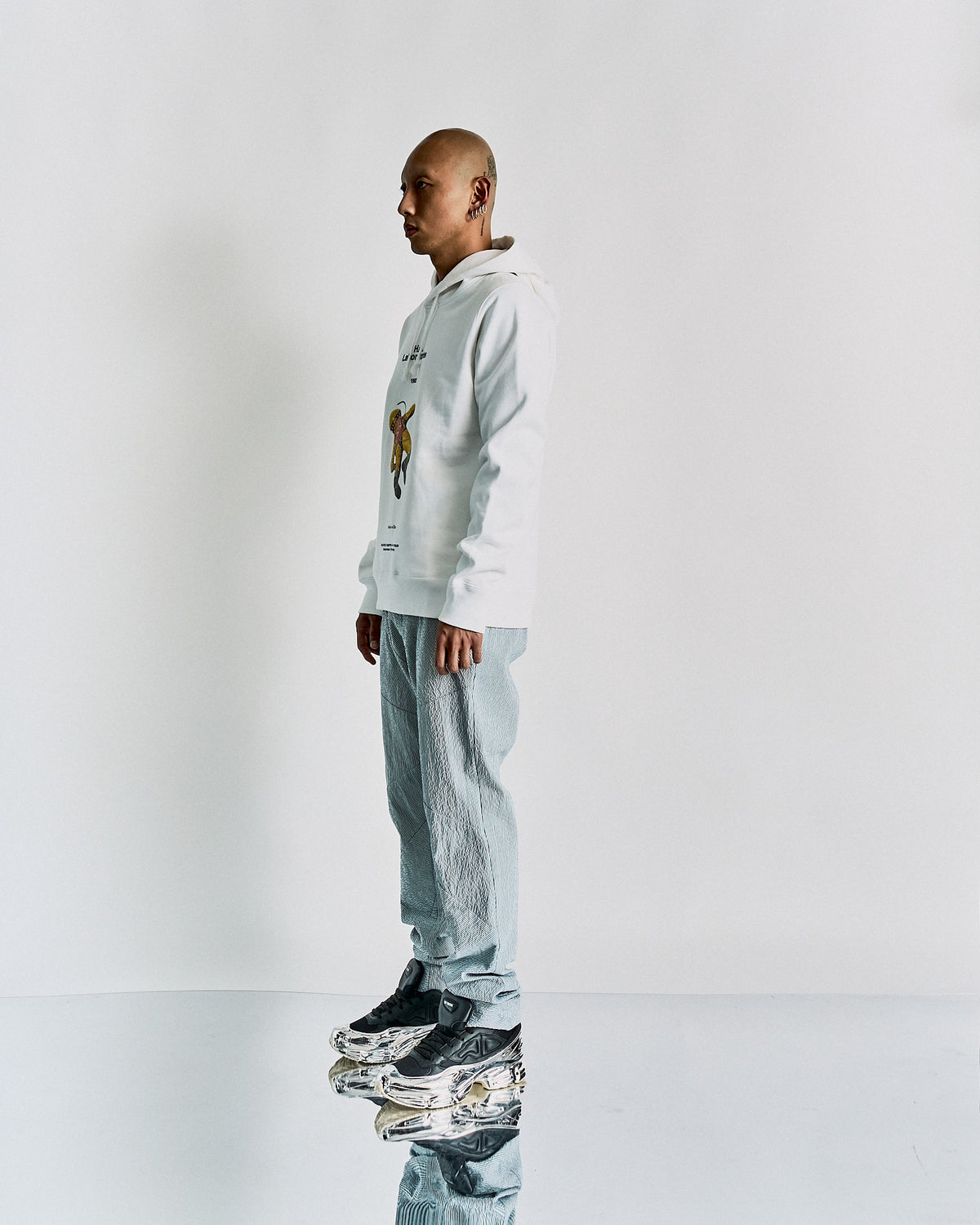 A person standing in a studio setting, wearing a white Undercover hoodie with a vintage-inspired 2001: A Space Odyssey print and gray pants.