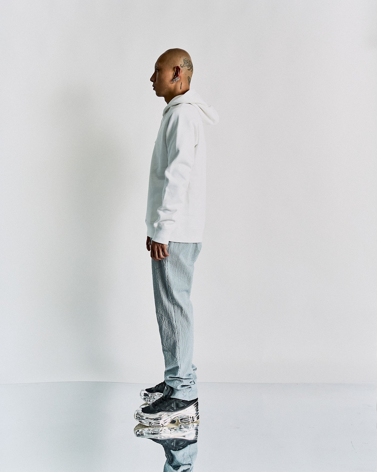 A person in a white Undercover hoodie and textured pants stands in profile on a reflective surface. 