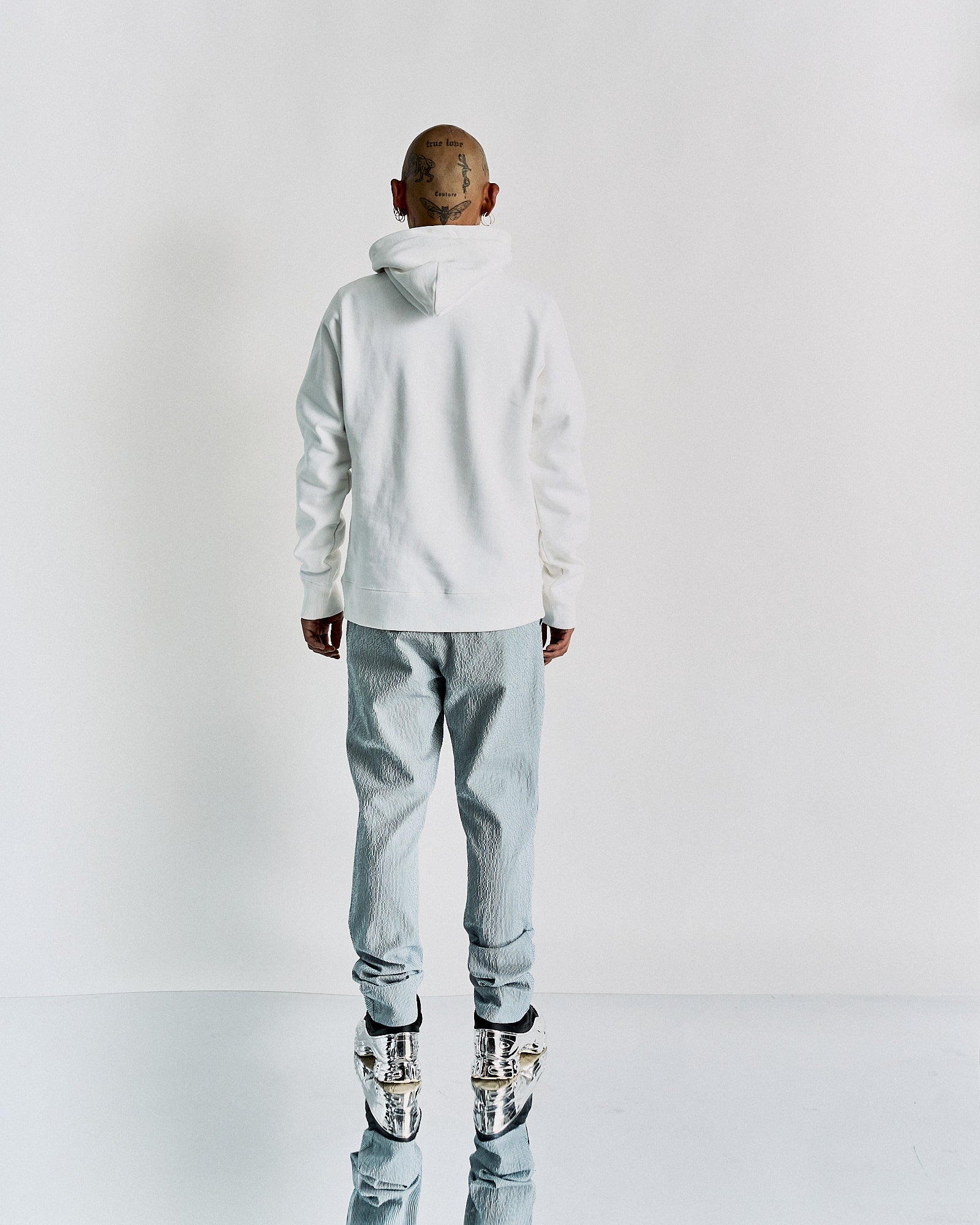 A person with a shaved head and tattoos stands facing away, wearing a white Undercover hoodie and silvery pants with reflective shoes, set against a plain backdrop.