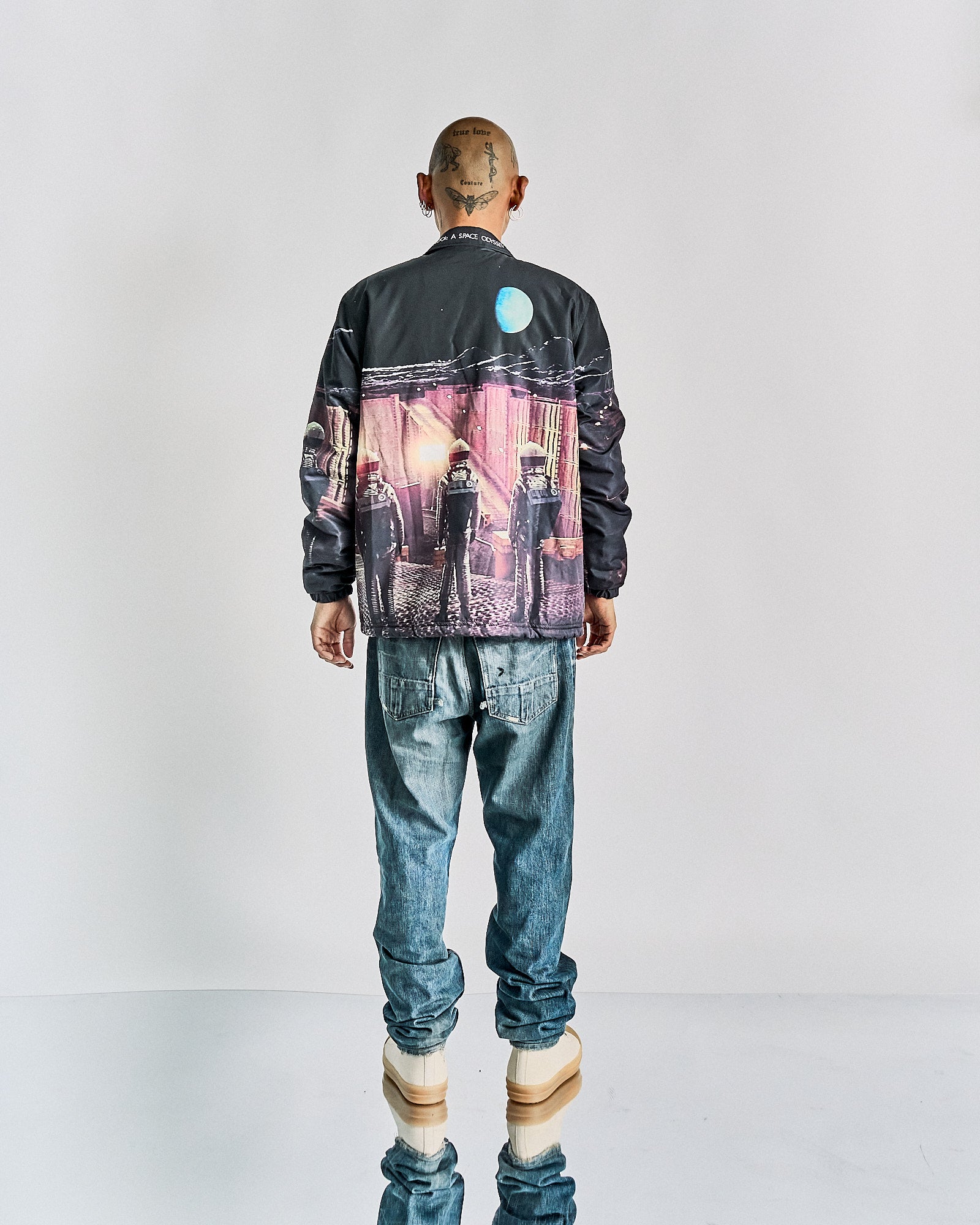 Bald person wearing an Undercover x Stanley Kubrick 2001: A Space Odyssey collaboration jacket with a space-themed scene showing astronauts, paired with blue jeans and beige platform shoes, standing against a plain backdrop.