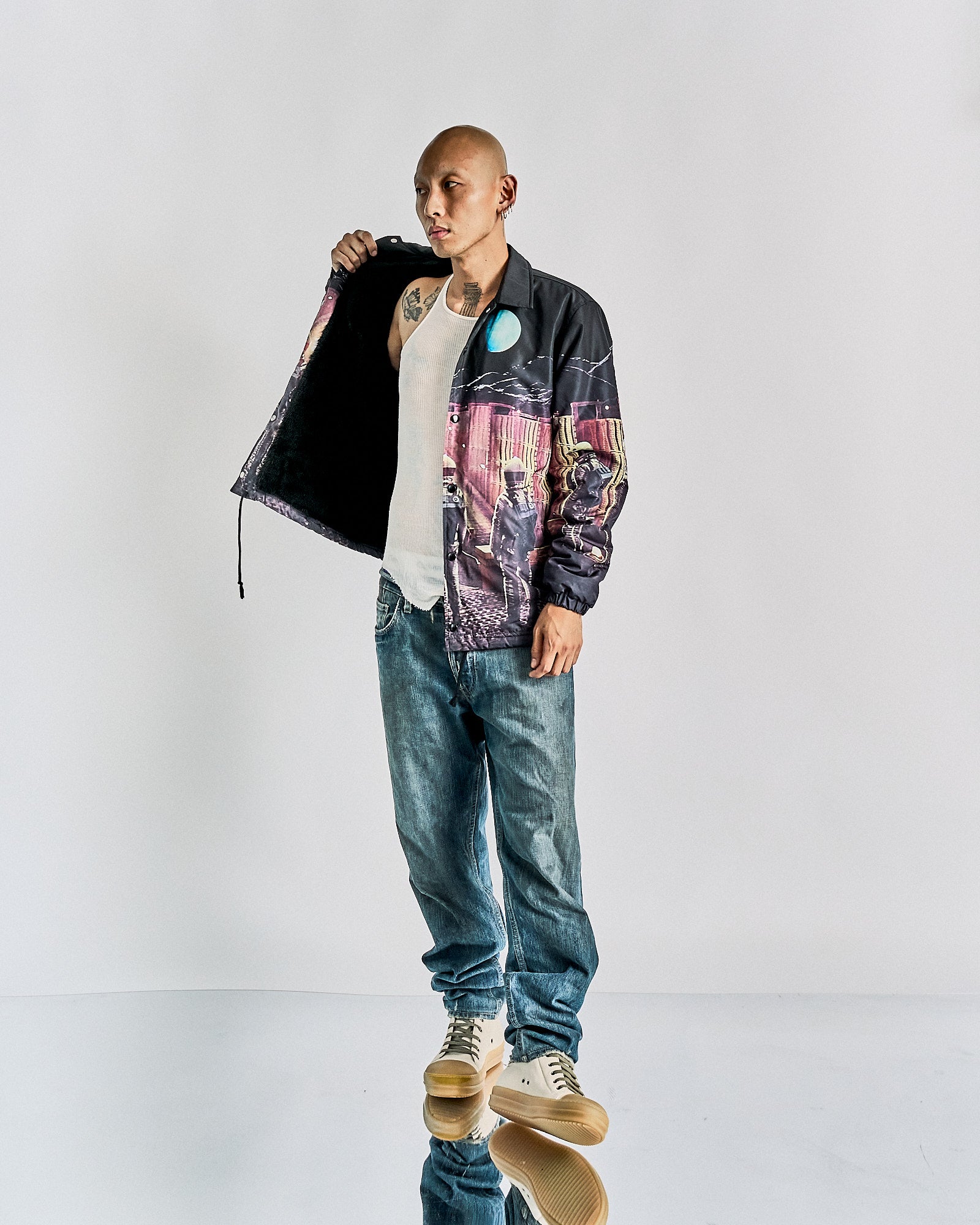 Bald man in a graphic Undercover x 2001: A Space Odyssey jacket and jeans stands confidently against a plain backdrop, displaying inner lining. He wears a white tank top and sneakers. Casual and bold.