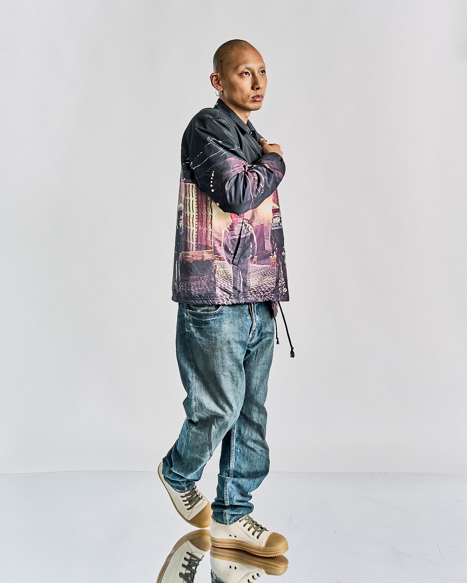 A person in casual attire stands confidently against a plain background. They're wearing a graphic Undercover x Stanley Kubrick jacket, loose denim jeans, and sneakers, exuding a relaxed style.