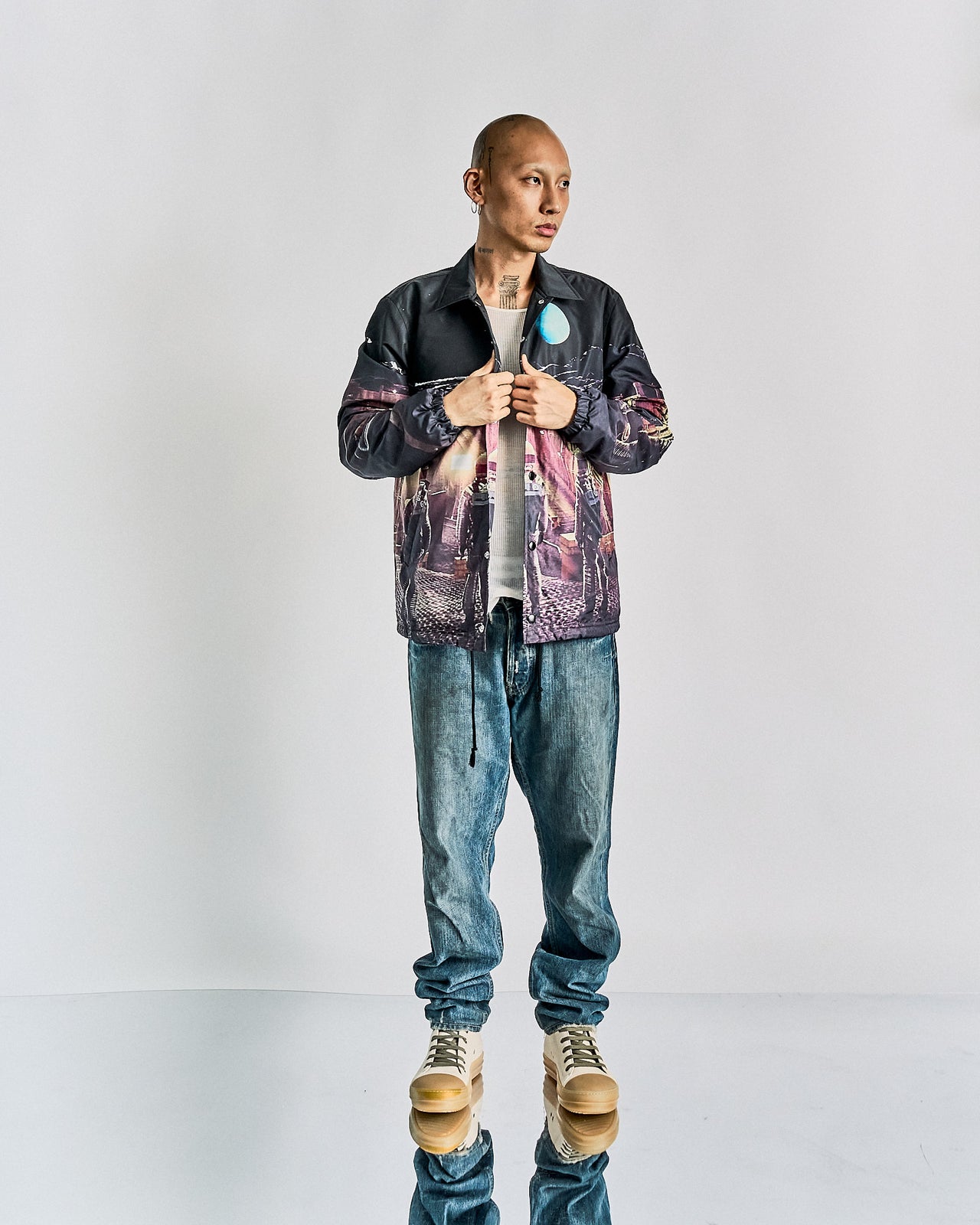 A person stands confidently in casual attire, wearing a printed Undercover x Stanley Kubrick jacket, white shirt, loose blue jeans, and beige high-top sneakers. The pose conveys a relaxed, stylish tone.