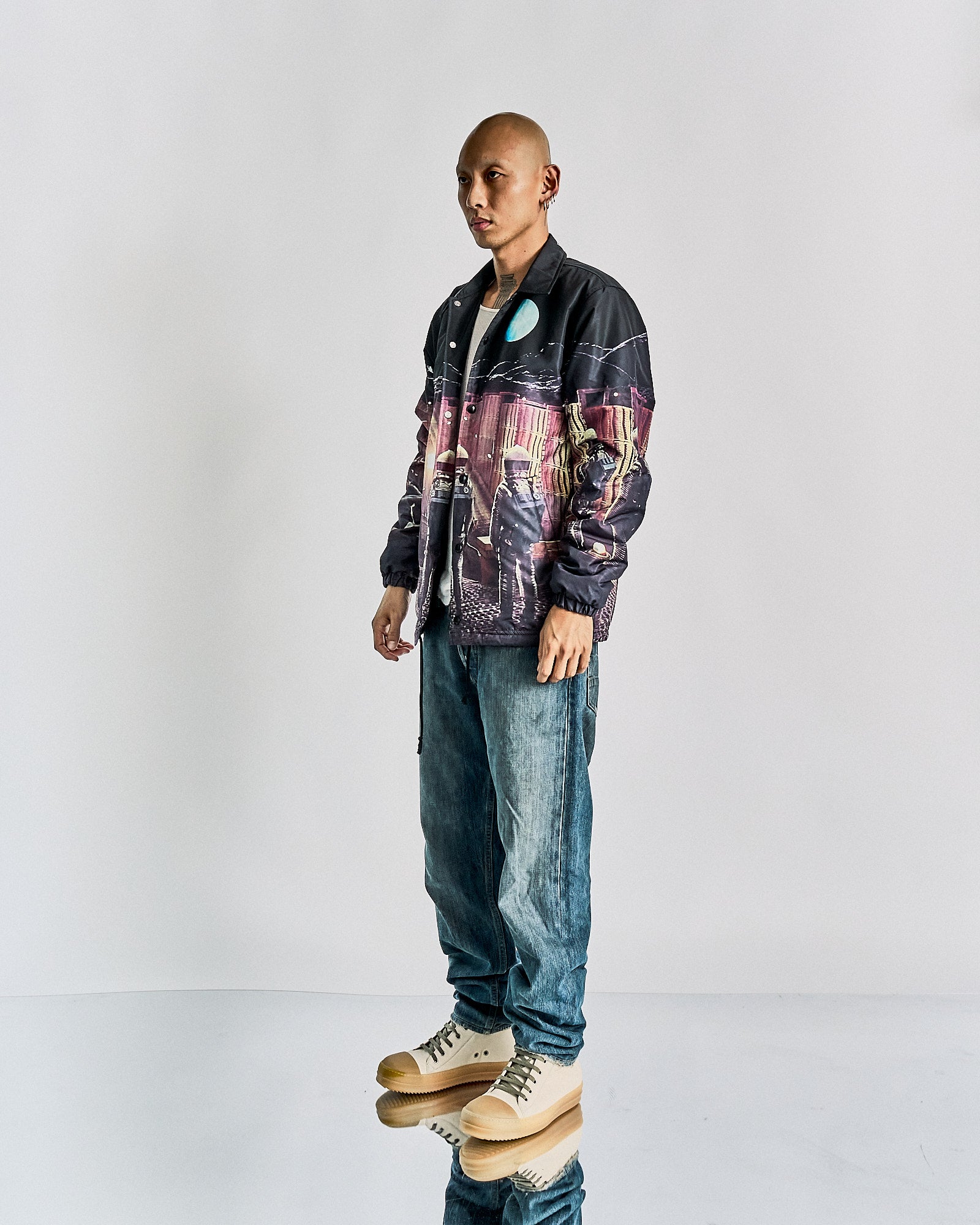 A person stands confidently in casual attire, wearing a printed Undercover x Stanley Kubrick jacket, white shirt, loose blue jeans, and beige high-top sneakers. 