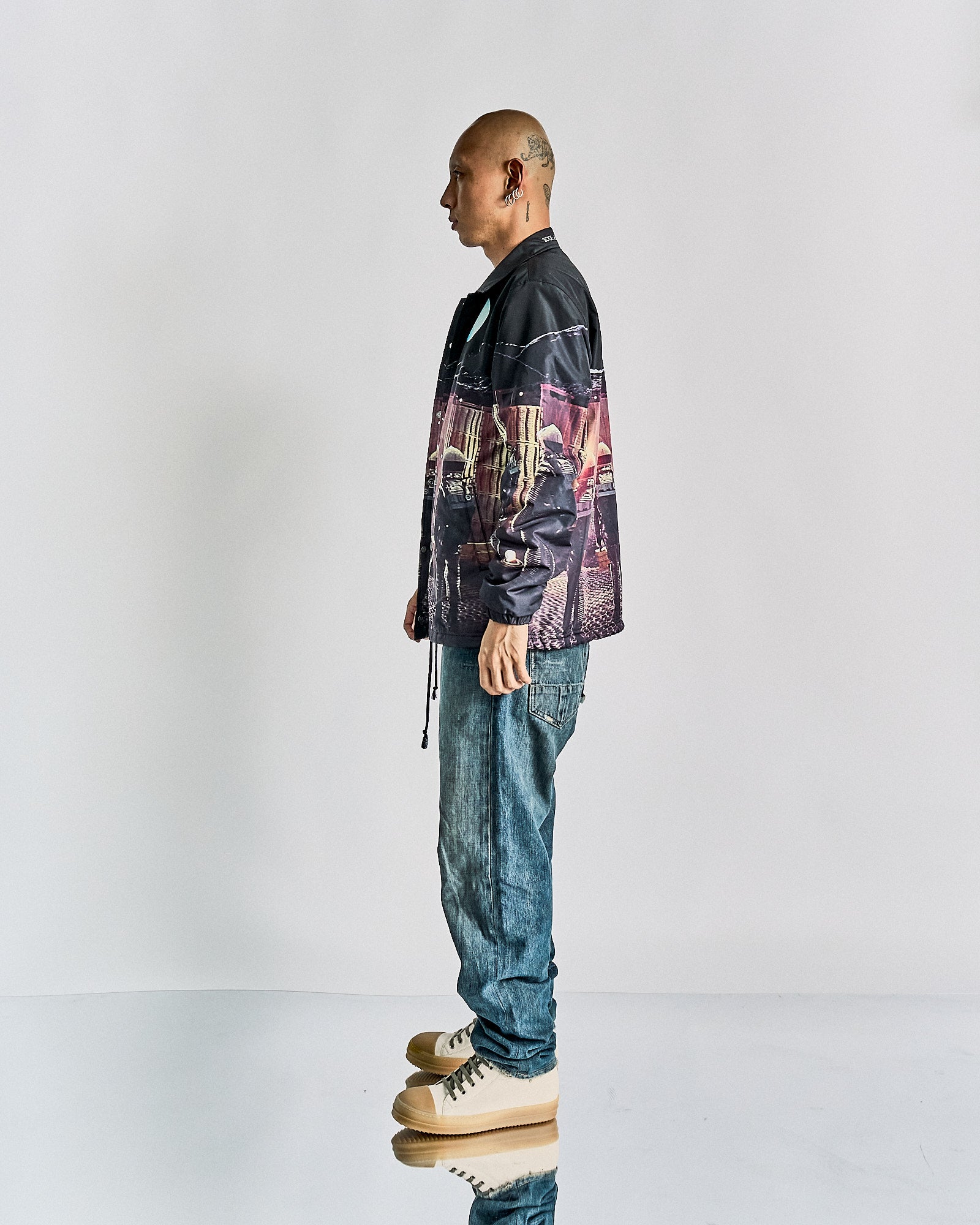 A person stands confidently in casual attire, wearing a printed Undercover x Stanley Kubrick jacket, white shirt, loose blue jeans, and beige high-top sneakers. The pose conveys a relaxed, stylish tone.