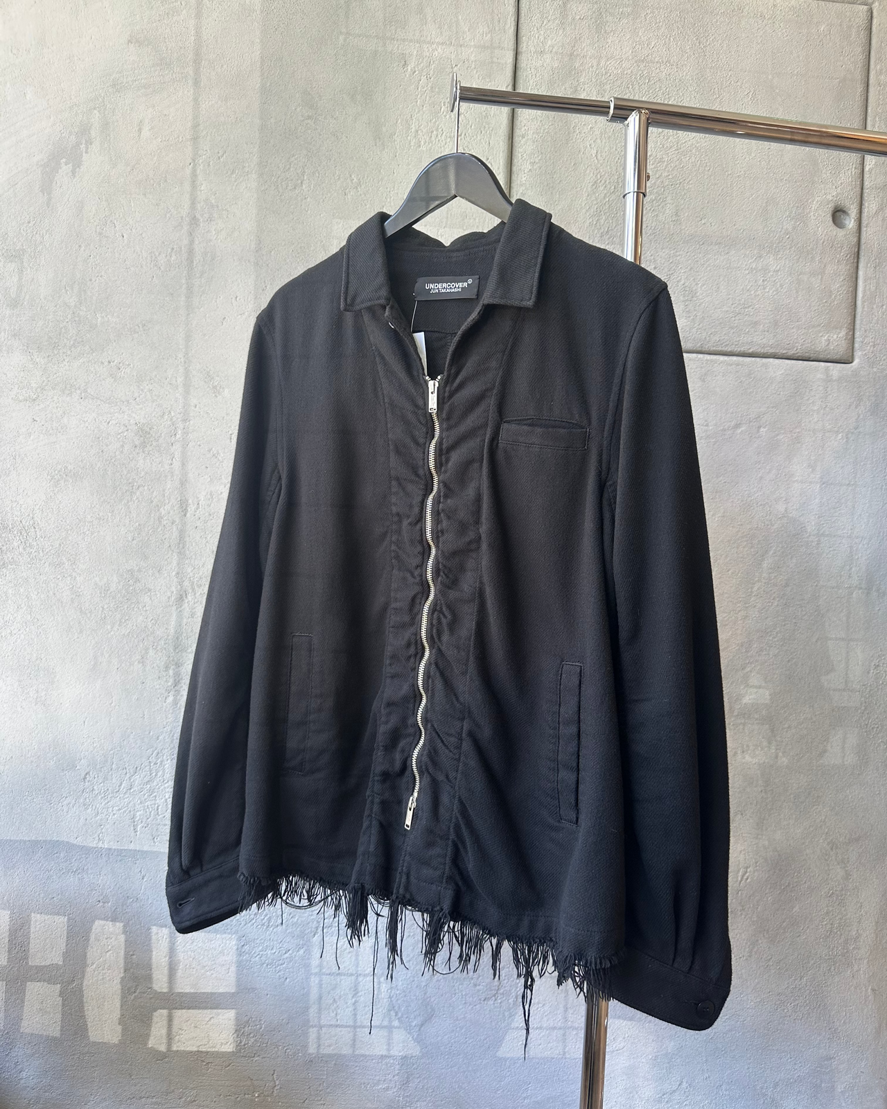 Black Undercover jacket with frayed edges on a hanger against a gray concrete wall.