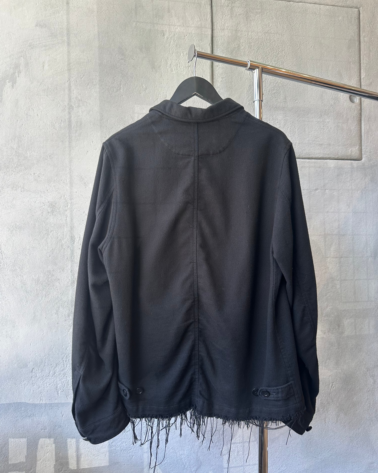 Black Undercover jacket with frayed edges on a hanger against a gray concrete wall.