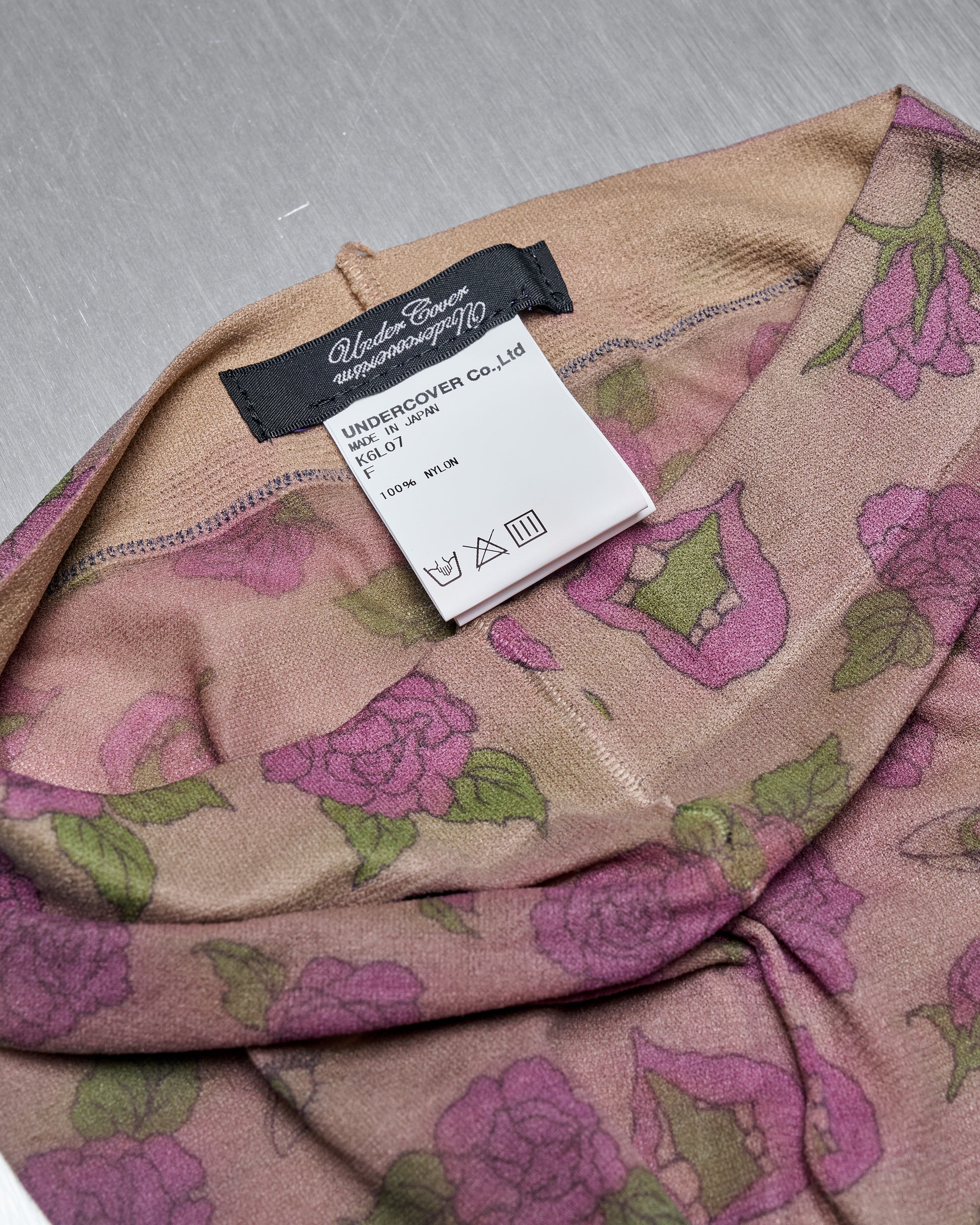 Floral-patterned beige garment with a label showing "Undercover Co. Ltd," made of nylon. The label also has care symbols. Background is a gray surface.