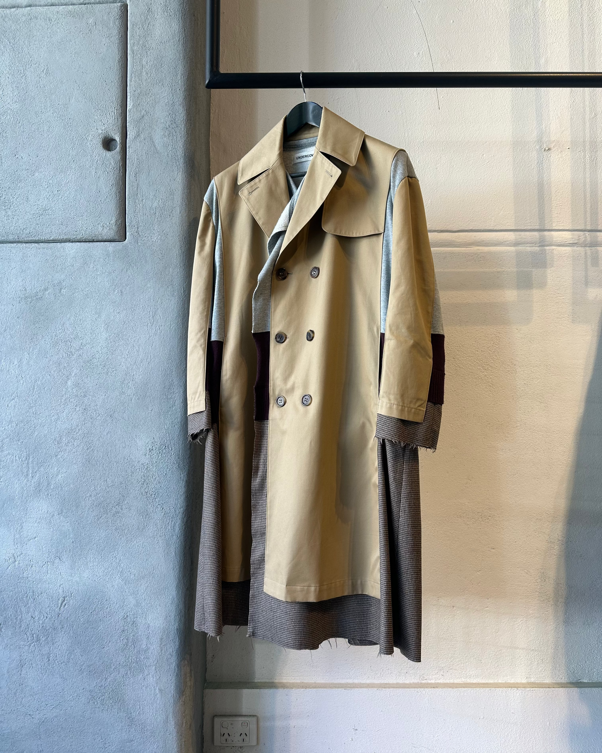 A creative, multi-layered Undercover trench coat hangs on a black hanger. It features a beige body with grey and brown patches, button details, and a textured finish.