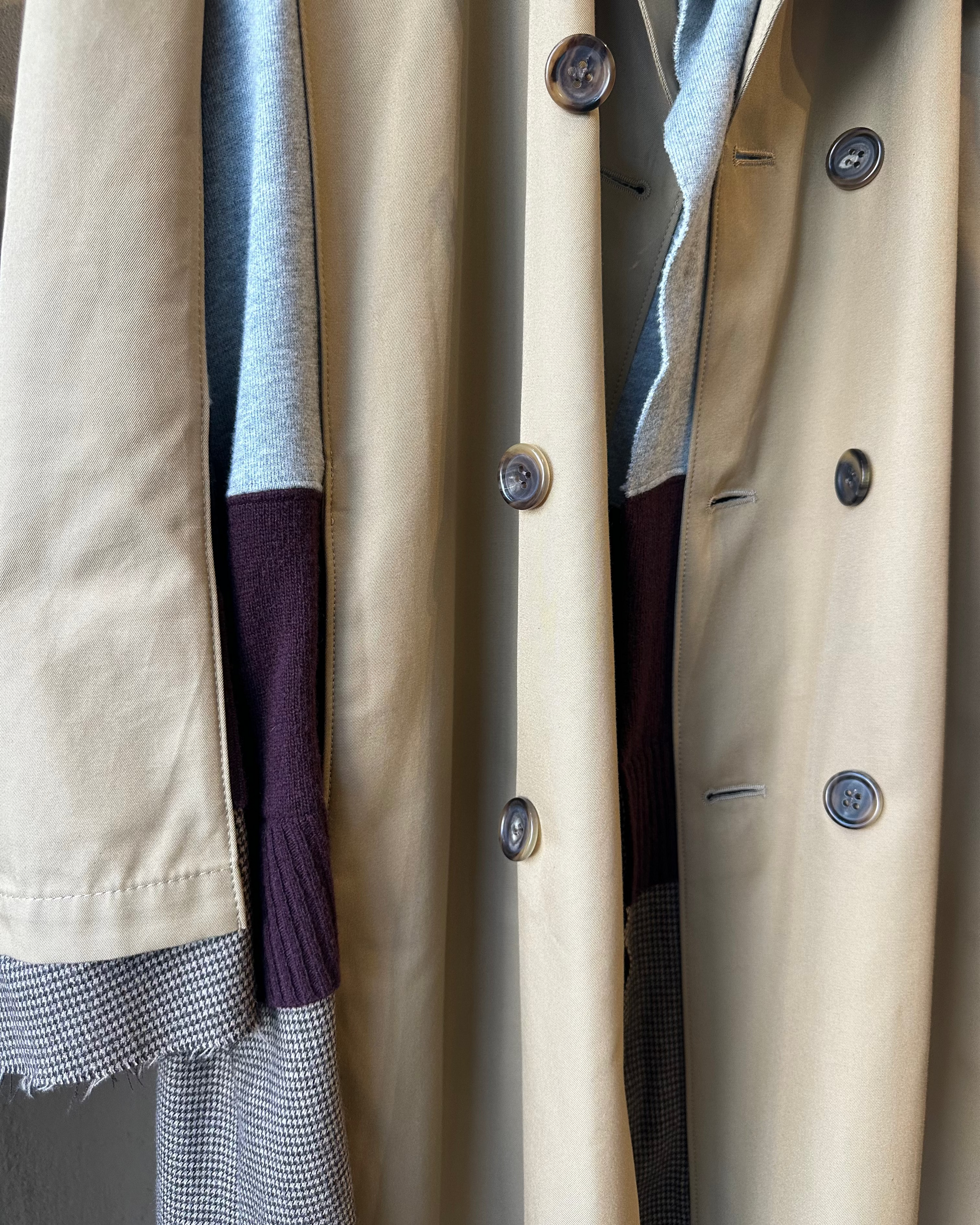 Close-up of a beige Undercover trench coat with buttons and a gray sweater peeking out.