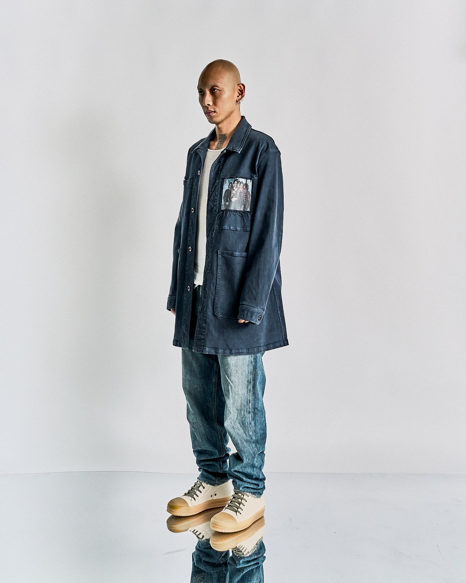 Man in a blue Undercover jacket with a photo patch, white shirt, and jeans. He stands on a reflective floor, wearing beige shoes, exuding a casual, laid-back vibe.