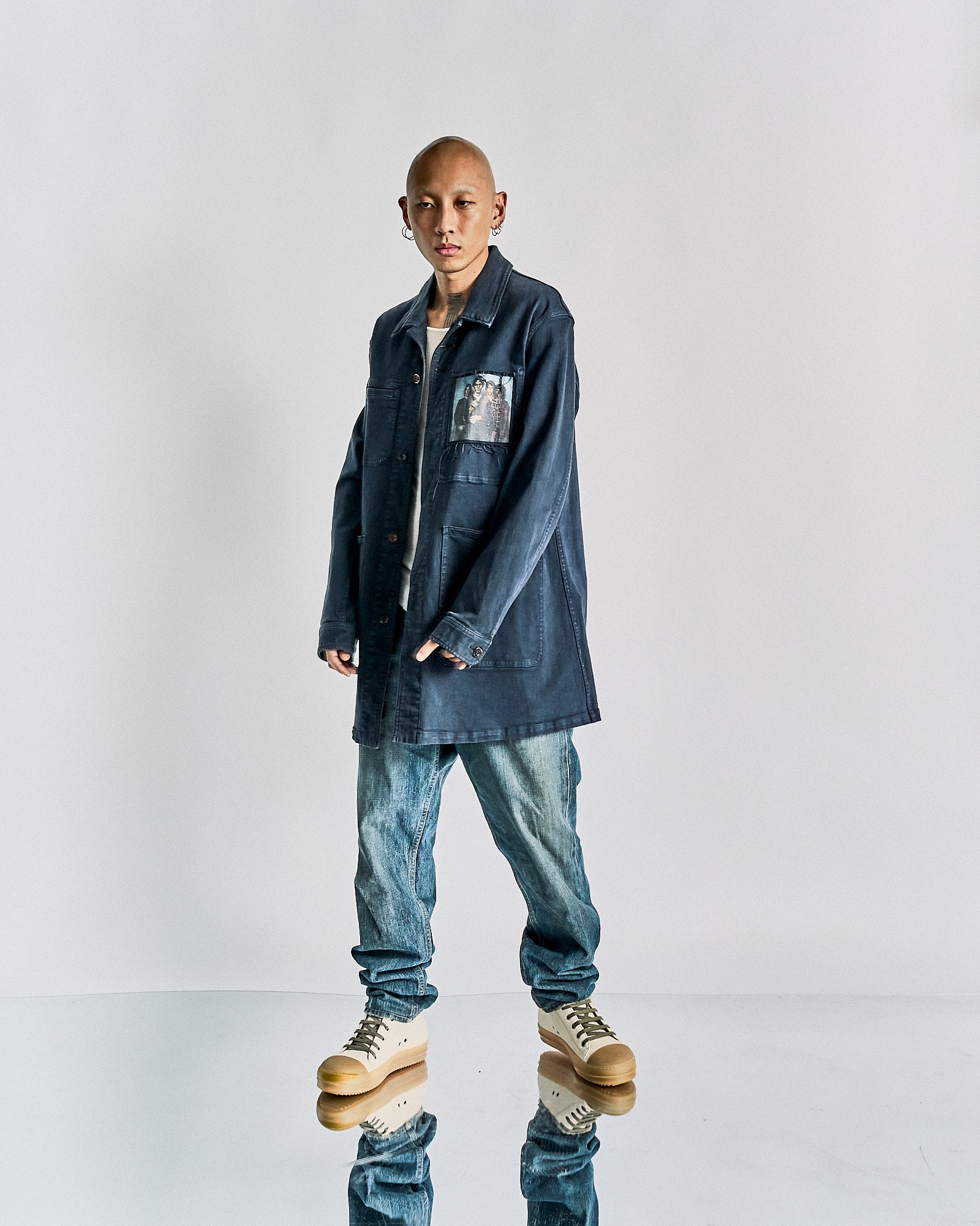 A person wears a long, dark denim Undercover jacket with a graphic patch, light blue jeans, and tan high-top sneakers, against a plain backdrop.