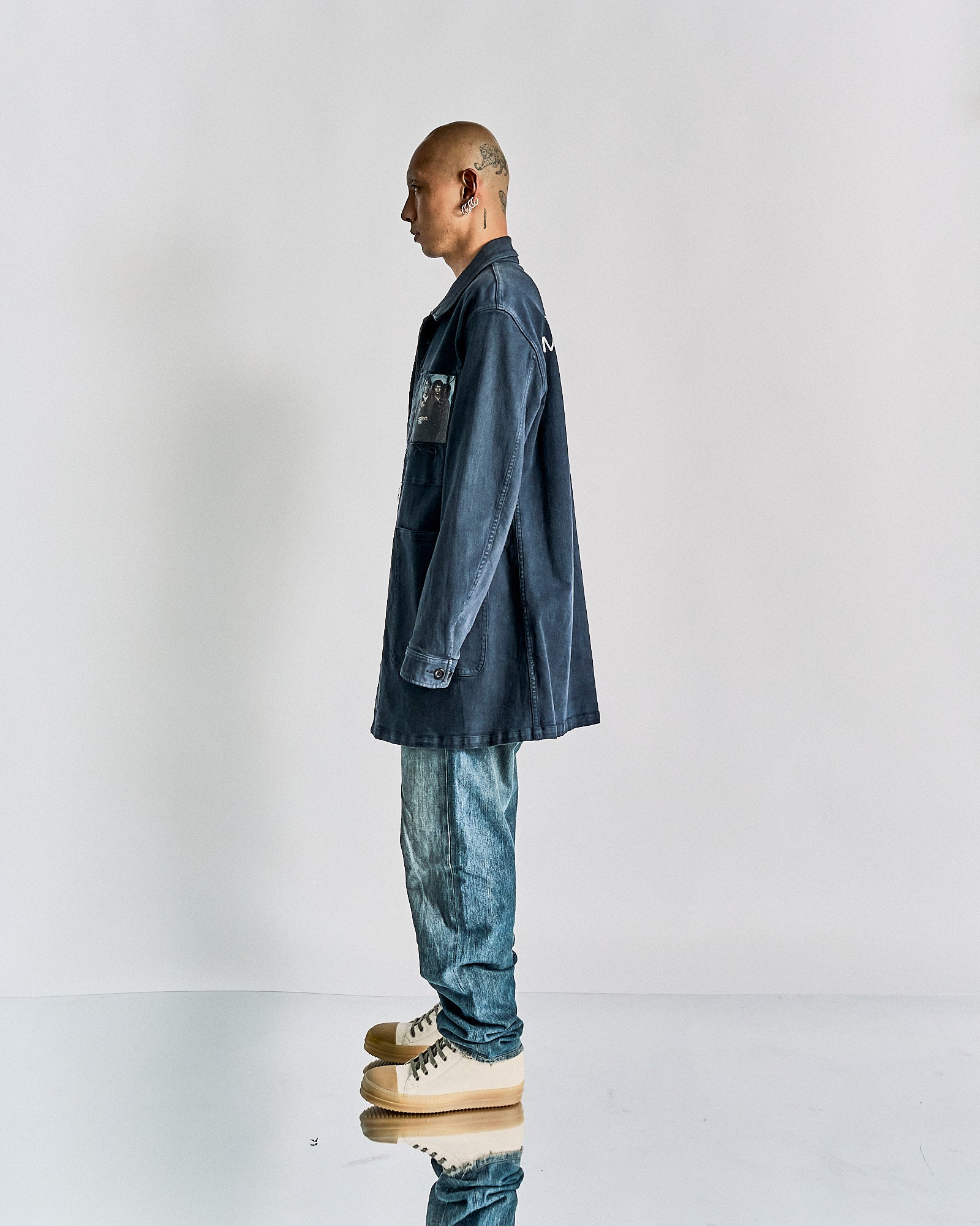 A person stands in profile against a plain backdrop, wearing a blue Undercover jacket with a patch, light blue jeans, and beige sneakers, exuding a casual, modern vibe.