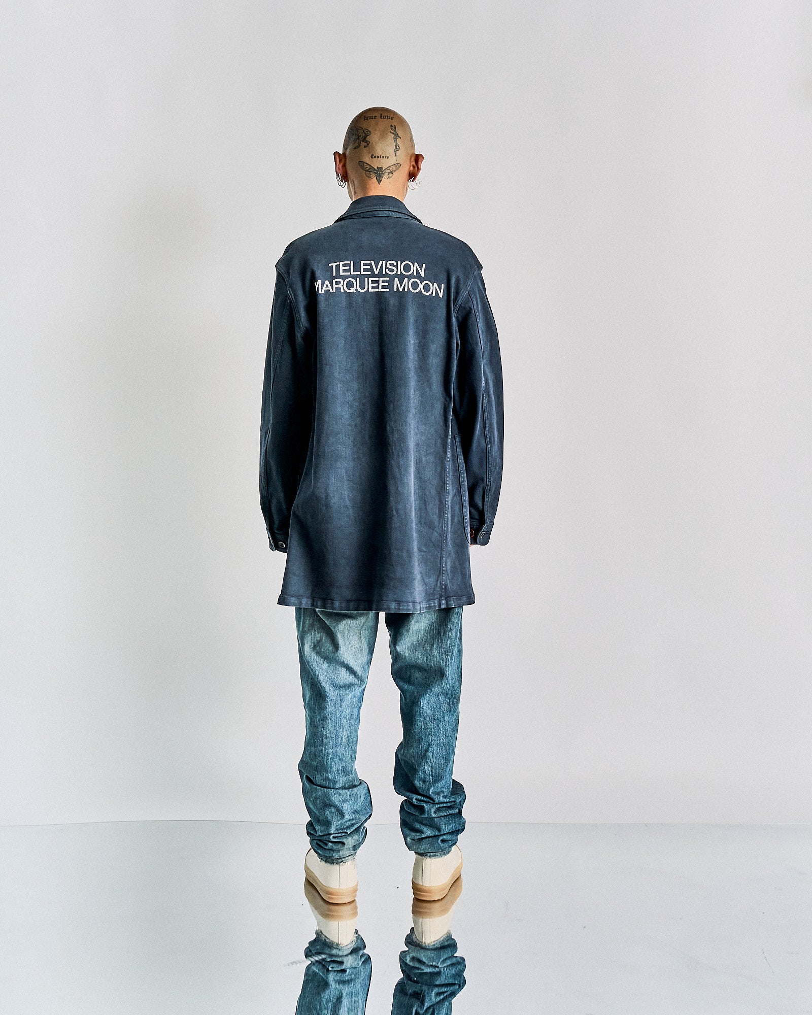 Man standing with back to the camera, wearing a long, dark denim Undercover shirt with "Television Marquee Moon" text, light jeans, and beige platform shoes. 