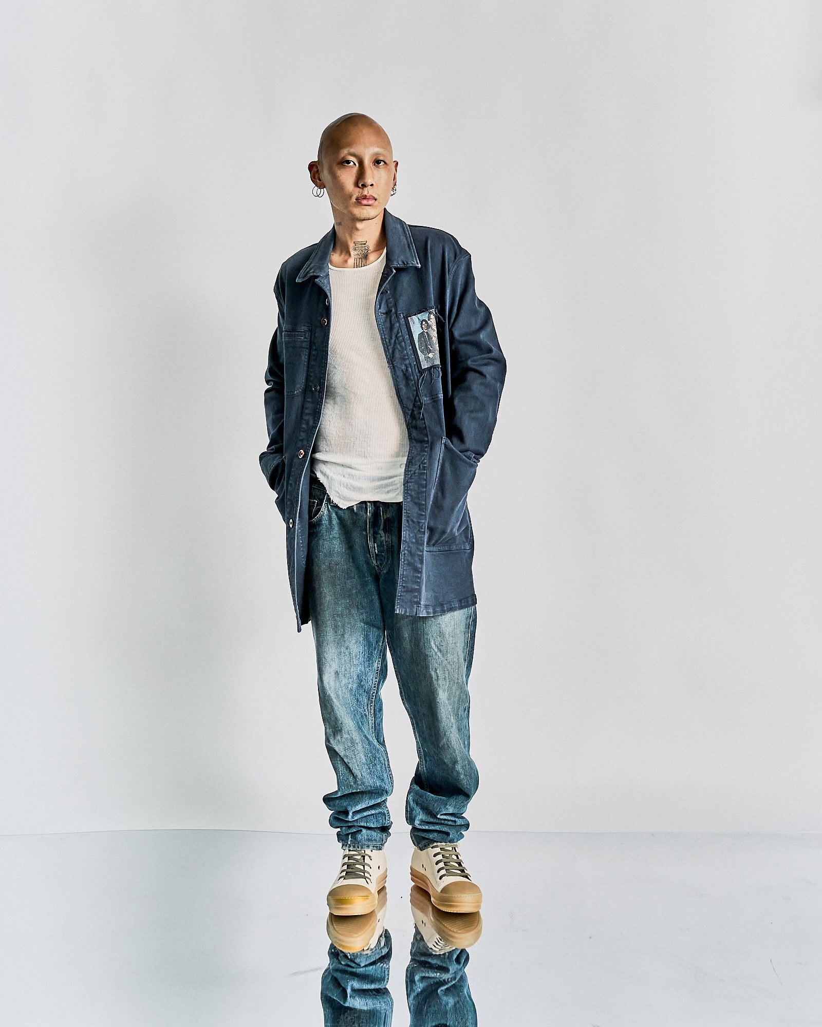 A person stands confidently against a plain backdrop, wearing a blue Undercover coat over a white shirt, jeans, and tan boots. 