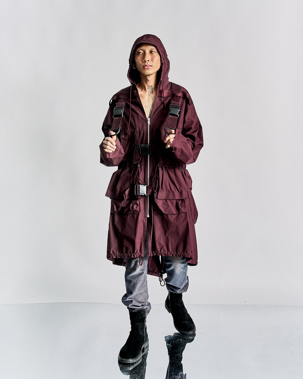 A person standing, wearing a purple Undercover hooded zip-up parka jacket with a backpack attached, in a studio setting against a white background.