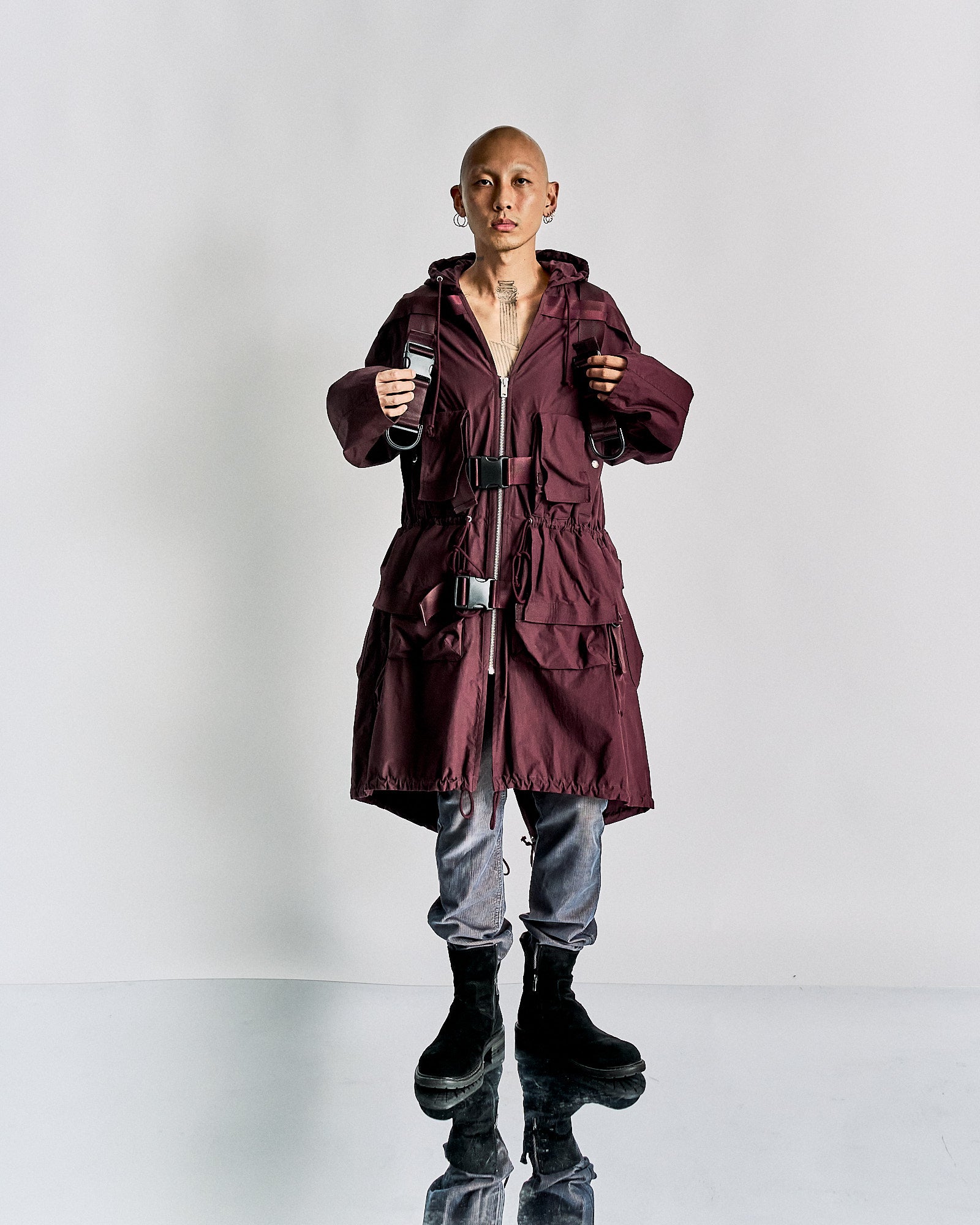 A person wearing a maroon, oversized Undercover trench coat with silver buckles and zippers. They have grey pants and black boots, standing confidently against a plain background.