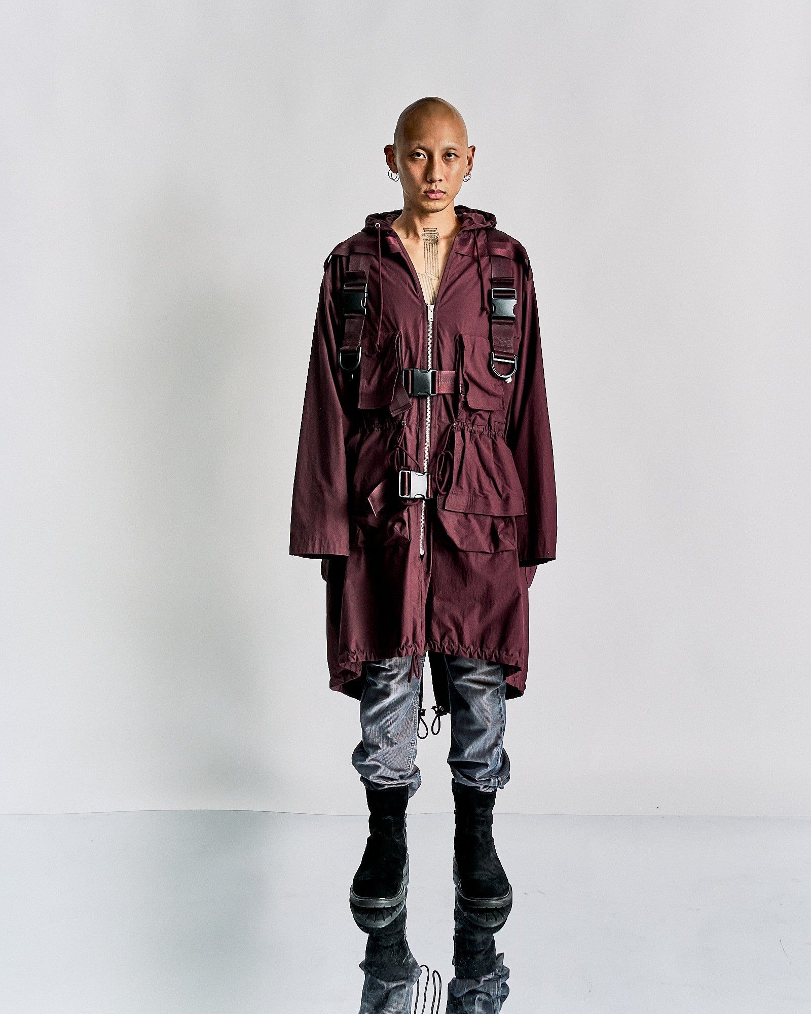 A person with a shaved head stands against a plain background, wearing a burgundy Undercover coat with buckle straps, grey pants, and black boots