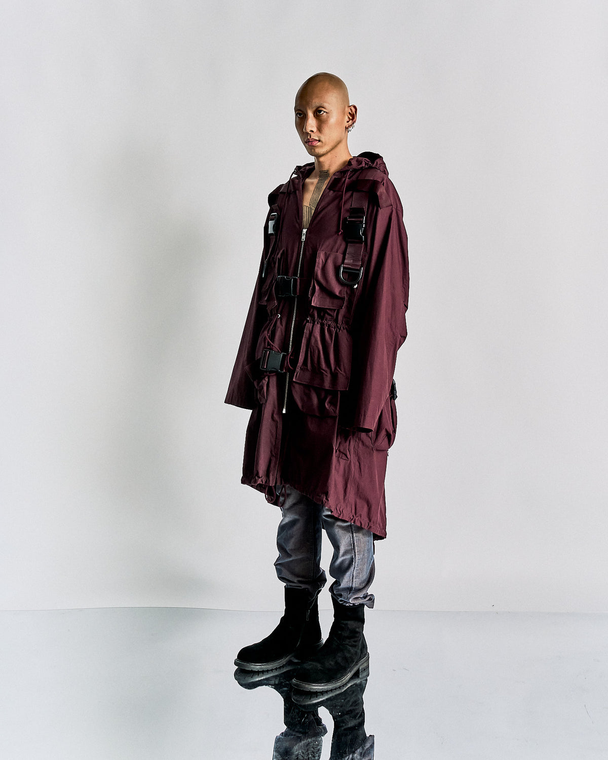 A model walks down the runway in a deep purple, Undercover layered outfit featuring a belted coat and cap, exuding a modern, edgy vibe. Dark boots complete the look.