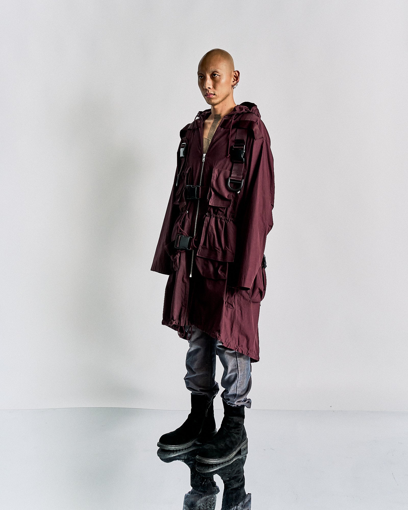 A person stands on a reflective surface, wearing an oversized maroon Undercover jacket with black buckles, grey jeans, and black boots. The expression is neutral.