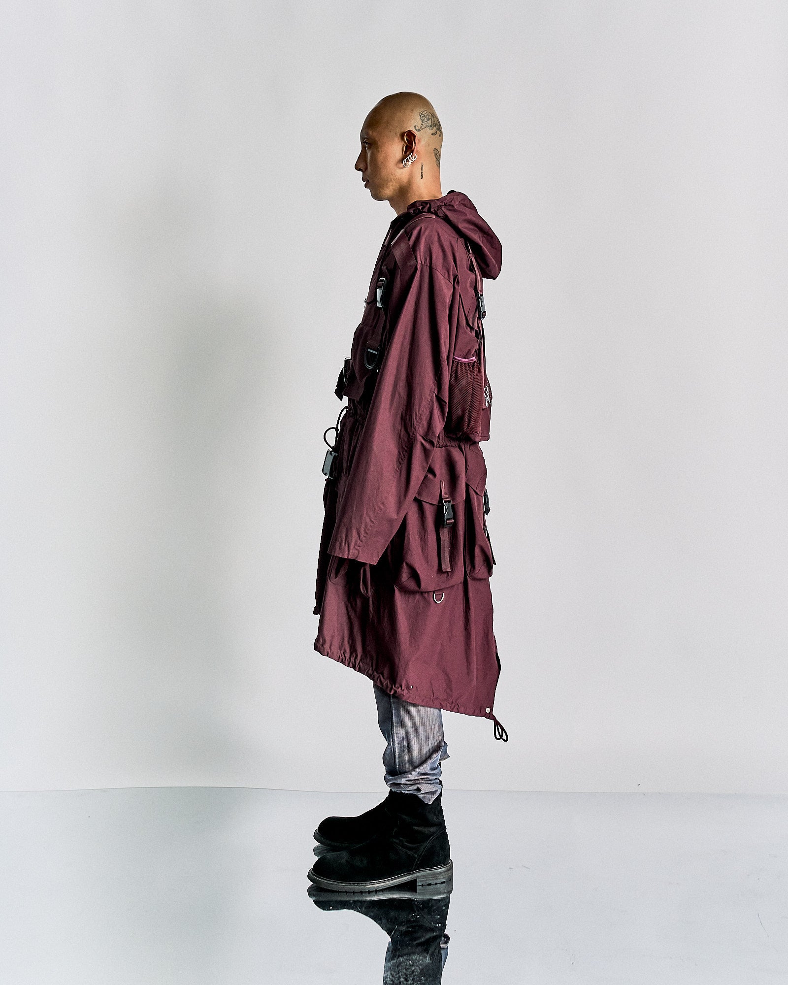 A person stands on a reflective surface, wearing an oversized maroon Undercover jacket with black buckles, grey jeans, and black boots. 
