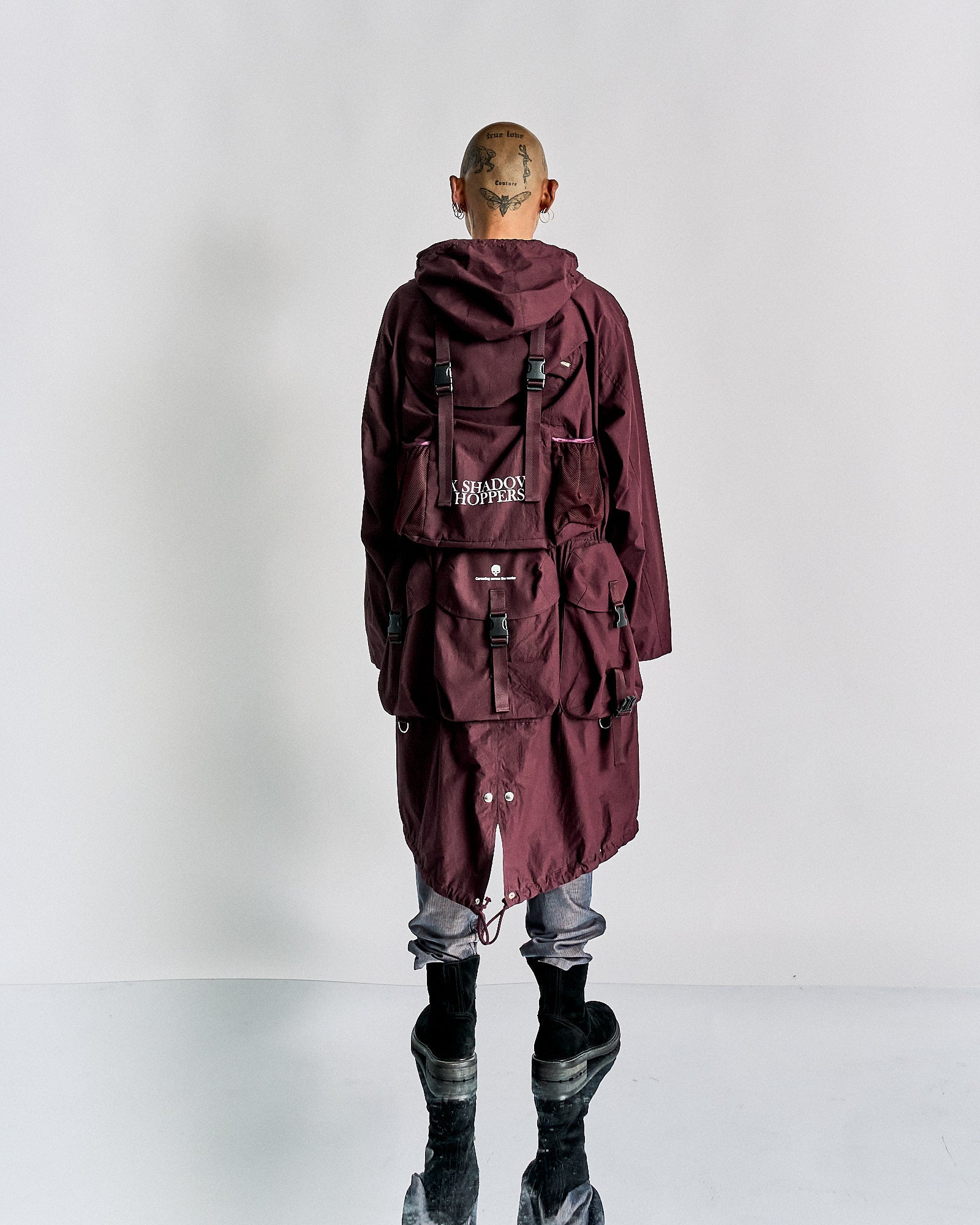 A person wearing a long Undercover brand maroon coat and a matching backpack with "Shadow Hoppers" text, standing against a grey background