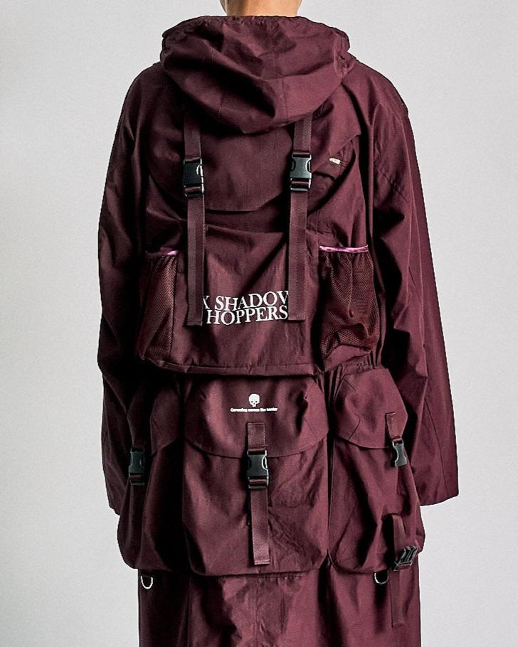A person wearing a burgundy hooded Undercover jacket with large pockets and black buckles on the back. The word "Shadow Hoppers" is printed in white.