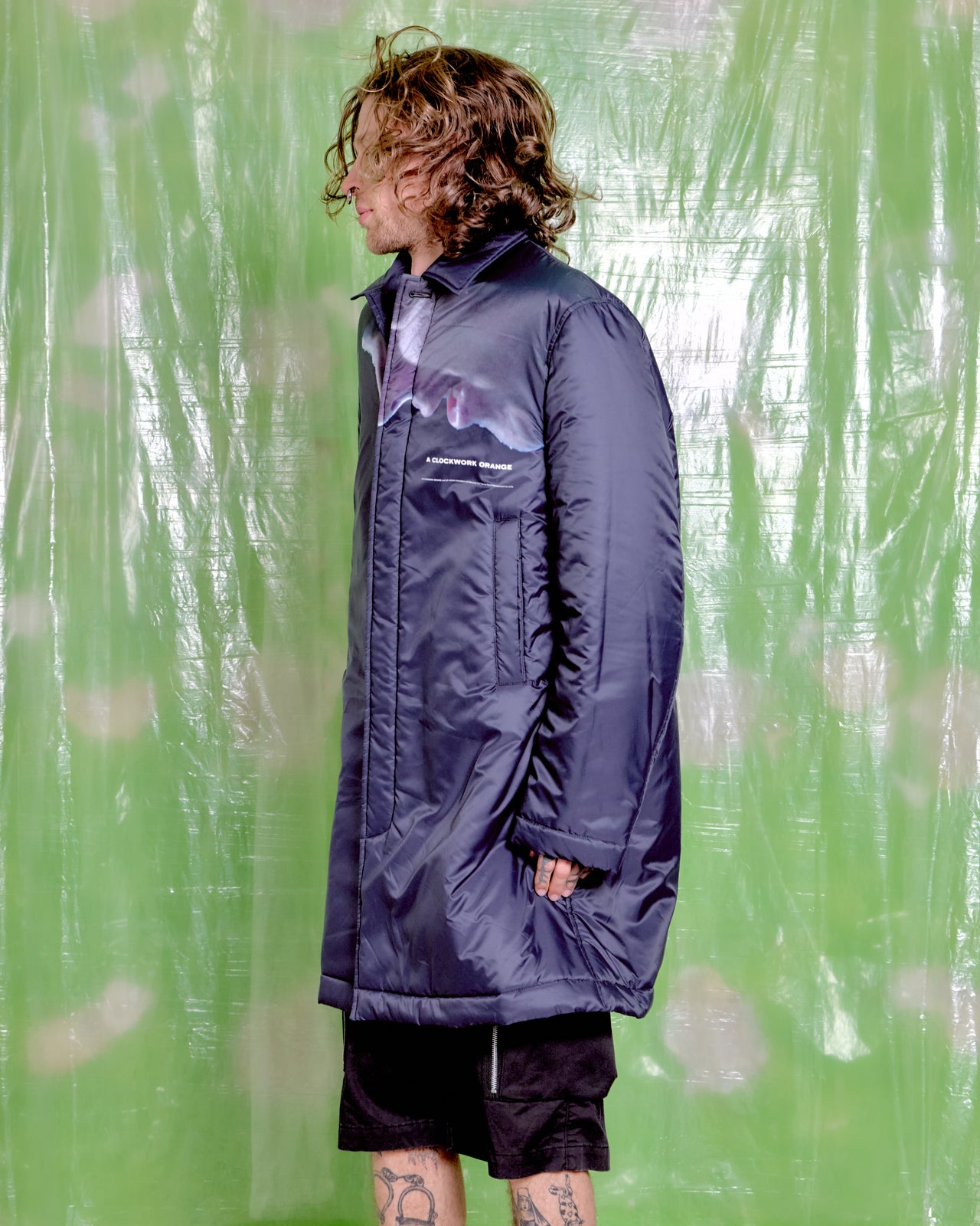Person wearing a long dark coat by Undercover X A Clockwork Orange with a visible brand logo against a green background.