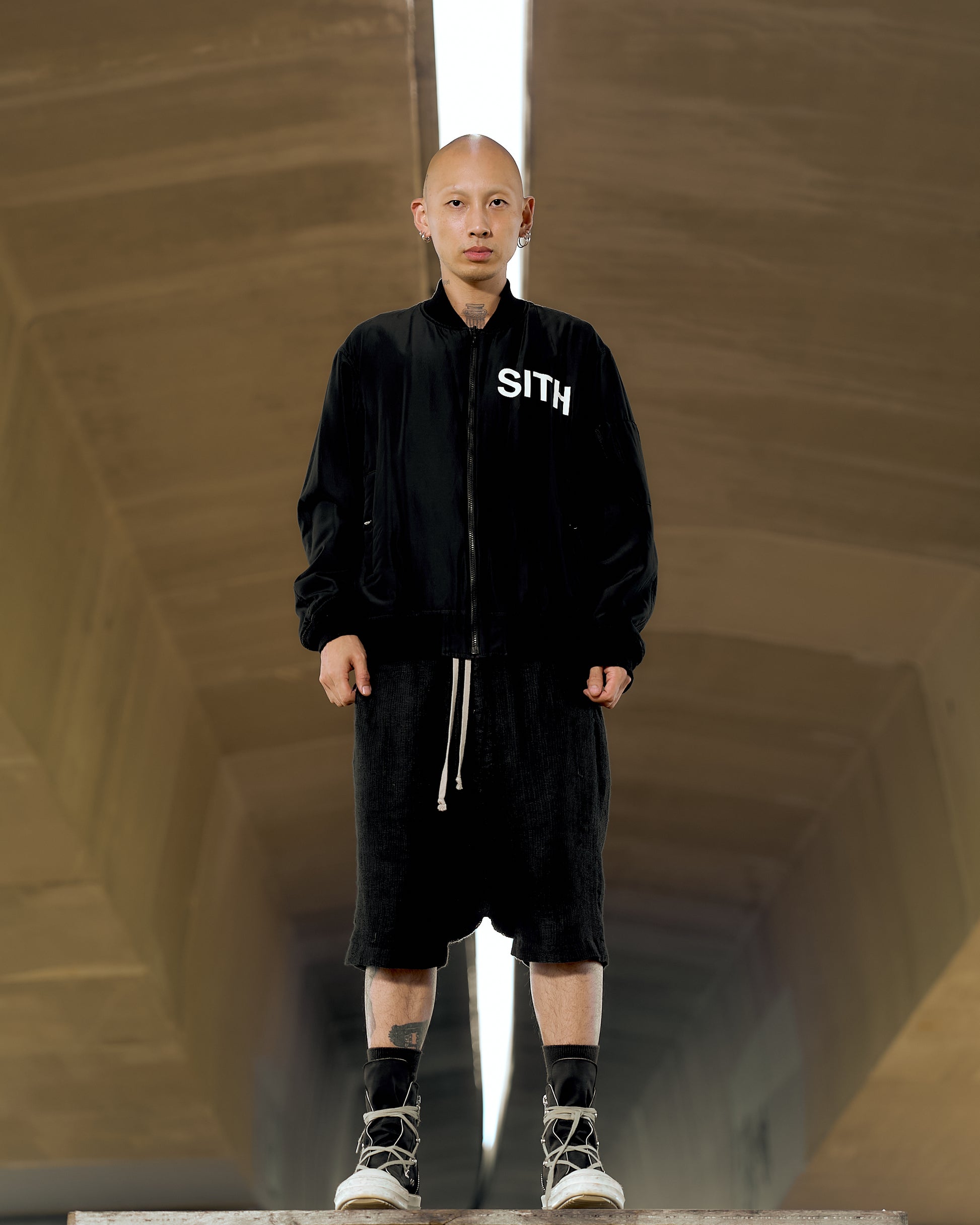 A person wearing a black reversible Undercover bomber jacket with 'SITH' written on the front, standing in a dimly lit space with geometric patterns in the background.