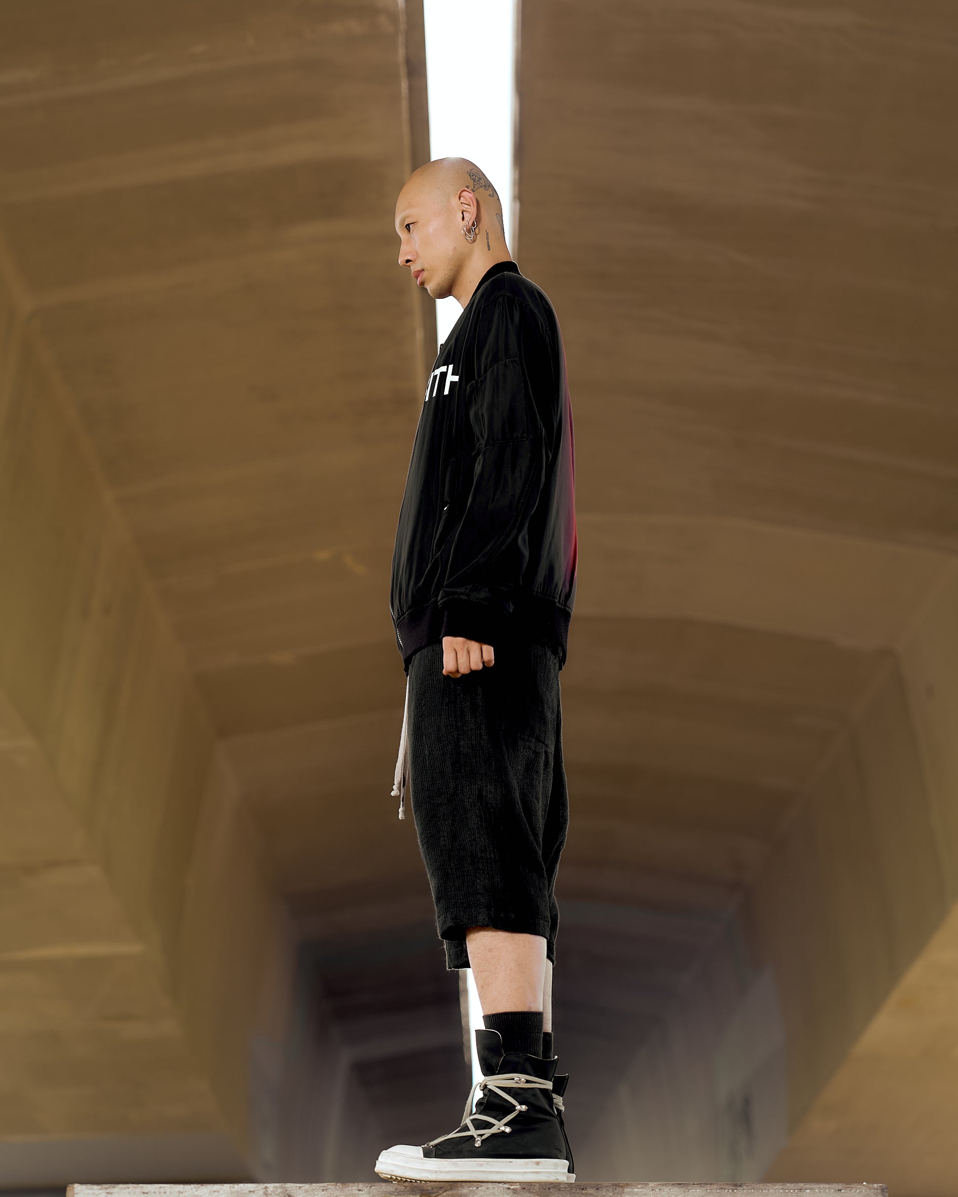 Bald person with earrings stands sideways in a tunnel wearing a black Undercover outfit and high-top sneakers, conveying a confident, edgy tone.
