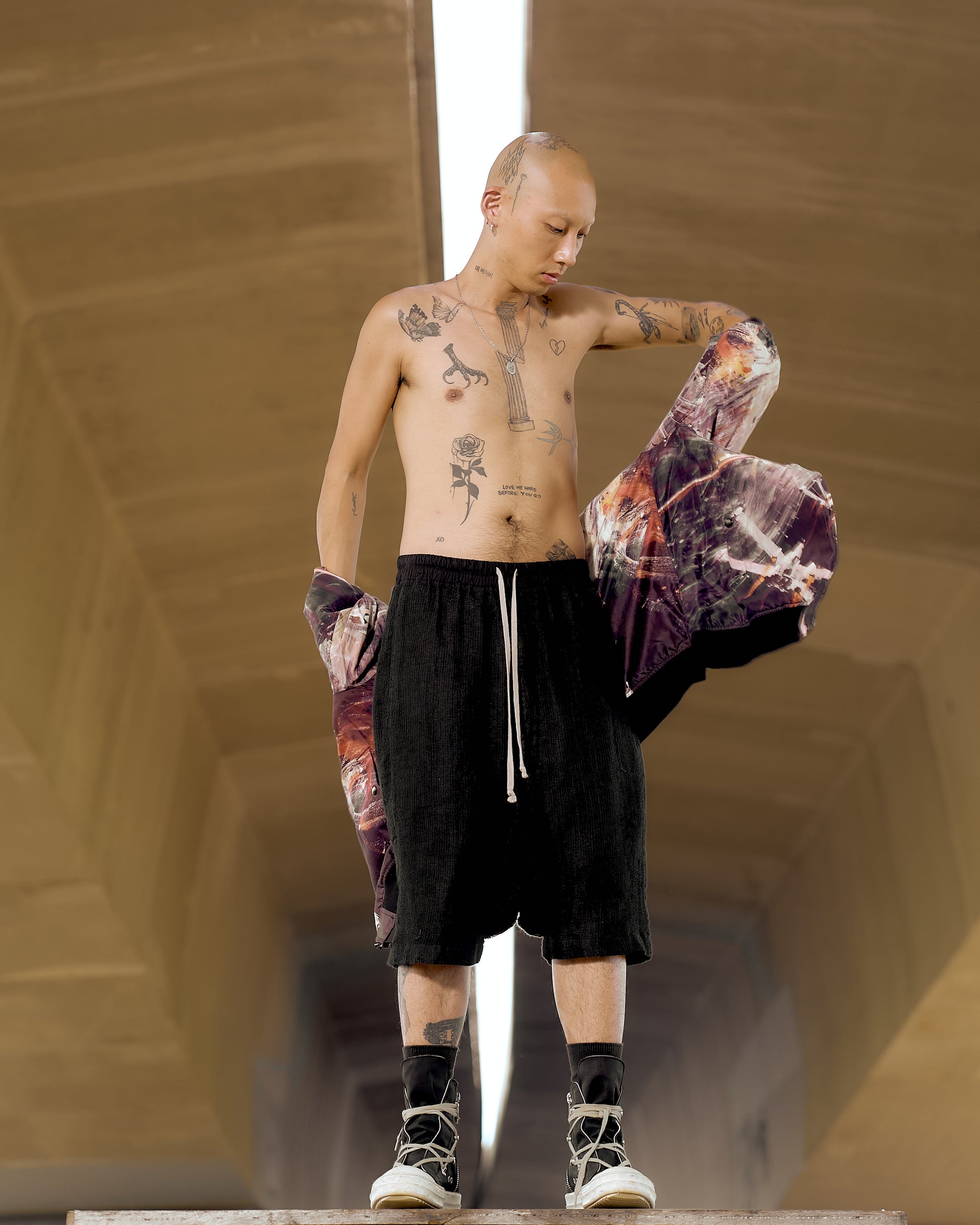 A bald, tattooed individual stands in an urban setting, wearing black shorts and sneakers, draping a colourful Undercover jacket over their shoulder, exuding confidence.