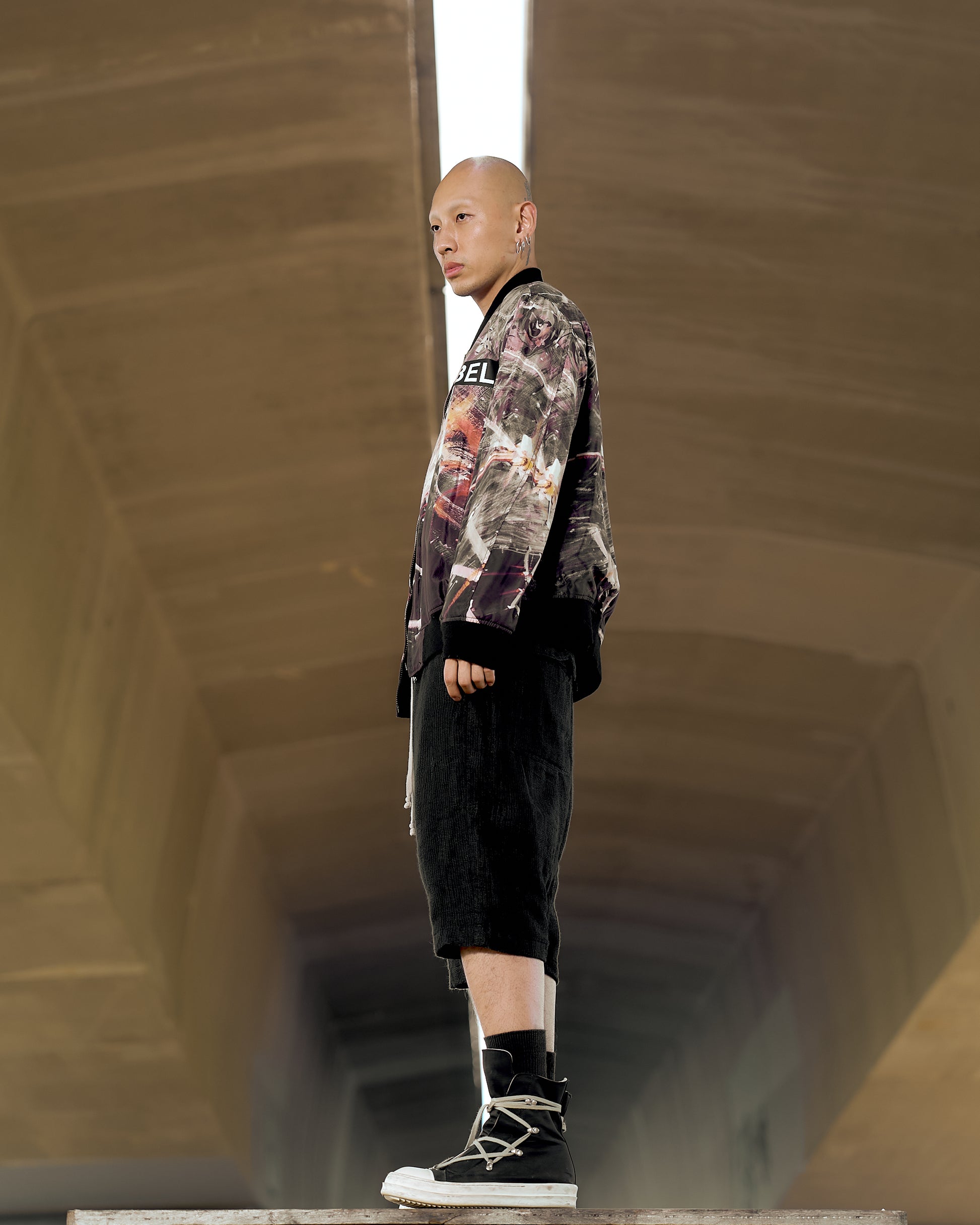 A man stands confidently in a tunnel wearing a patterned Undercover x Star Wars jacket, black shorts, and high-top sneakers. The lighting creates a dramatic and edgy mood.