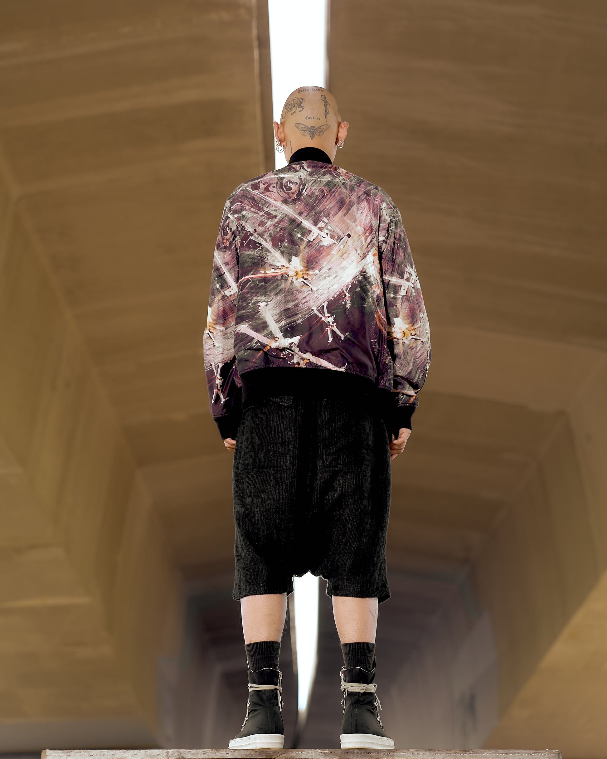 A person with a tattooed head is standing in an architectural setting, wearing a colourful abstract jacket by Undercover x Star Wars, black shorts, and sneakers. The scene feels modern and edgy.