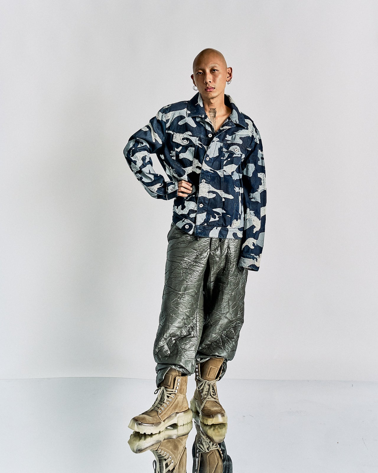 A person with a shaved head wears a blue camouflage Valentino jacket, metallic textured pants, and tan boots, standing confidently against a plain backdrop.